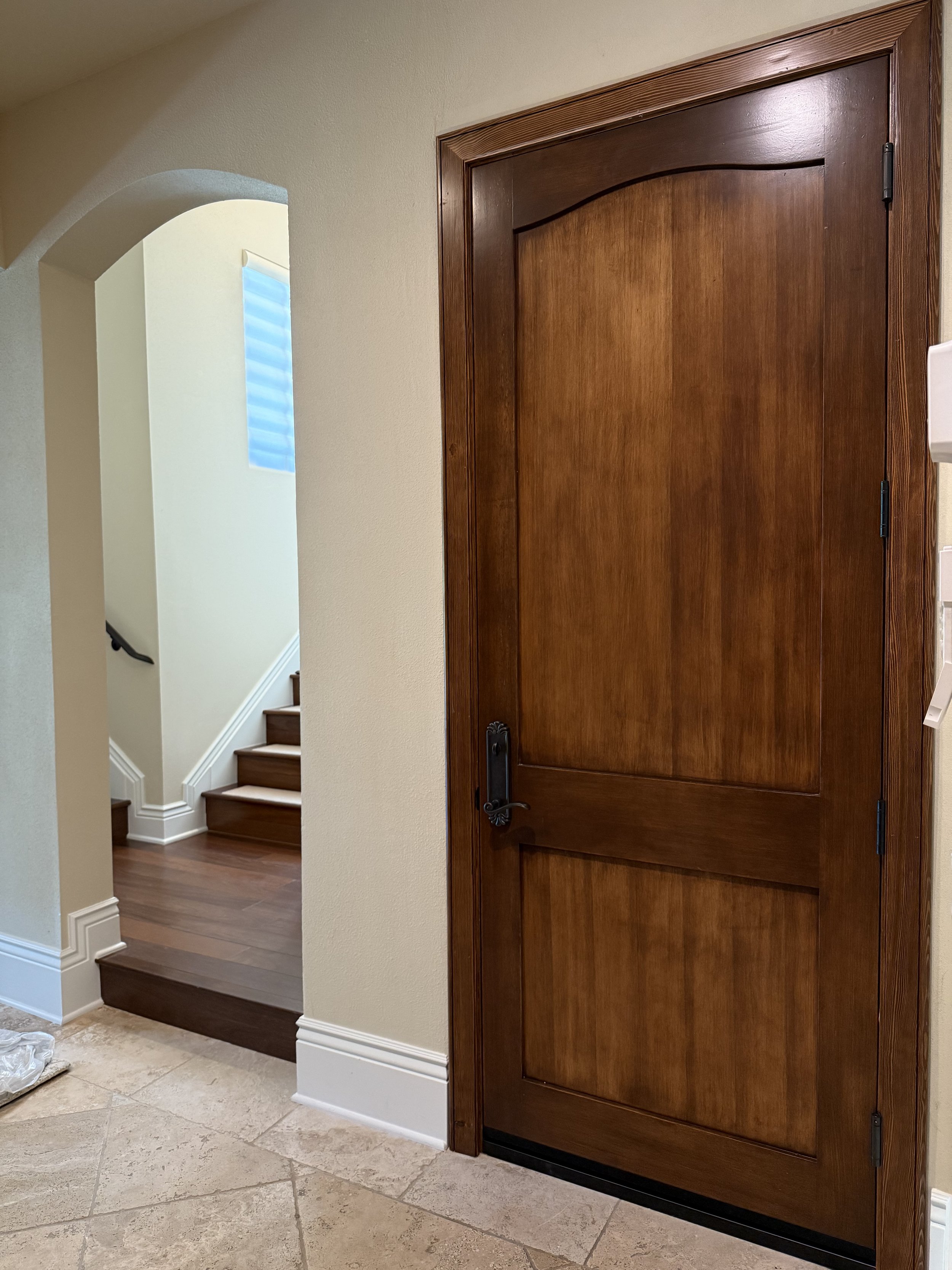 Interior view of a house featuring a closed wooden door, a beige wall, and a staircase with wooden steps and a black handrail near a small window with blinds.