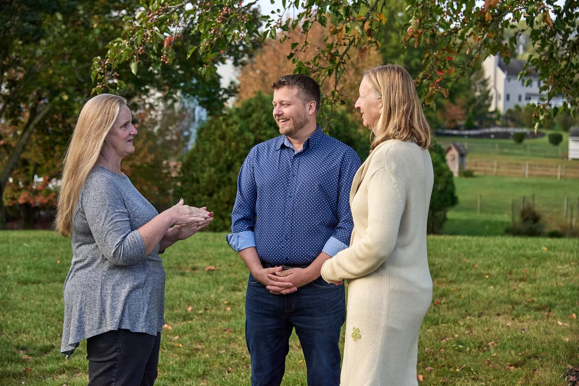 Chris and two others standing outdoors on a grassy area, engaged in conversation, with trees and houses in the background.