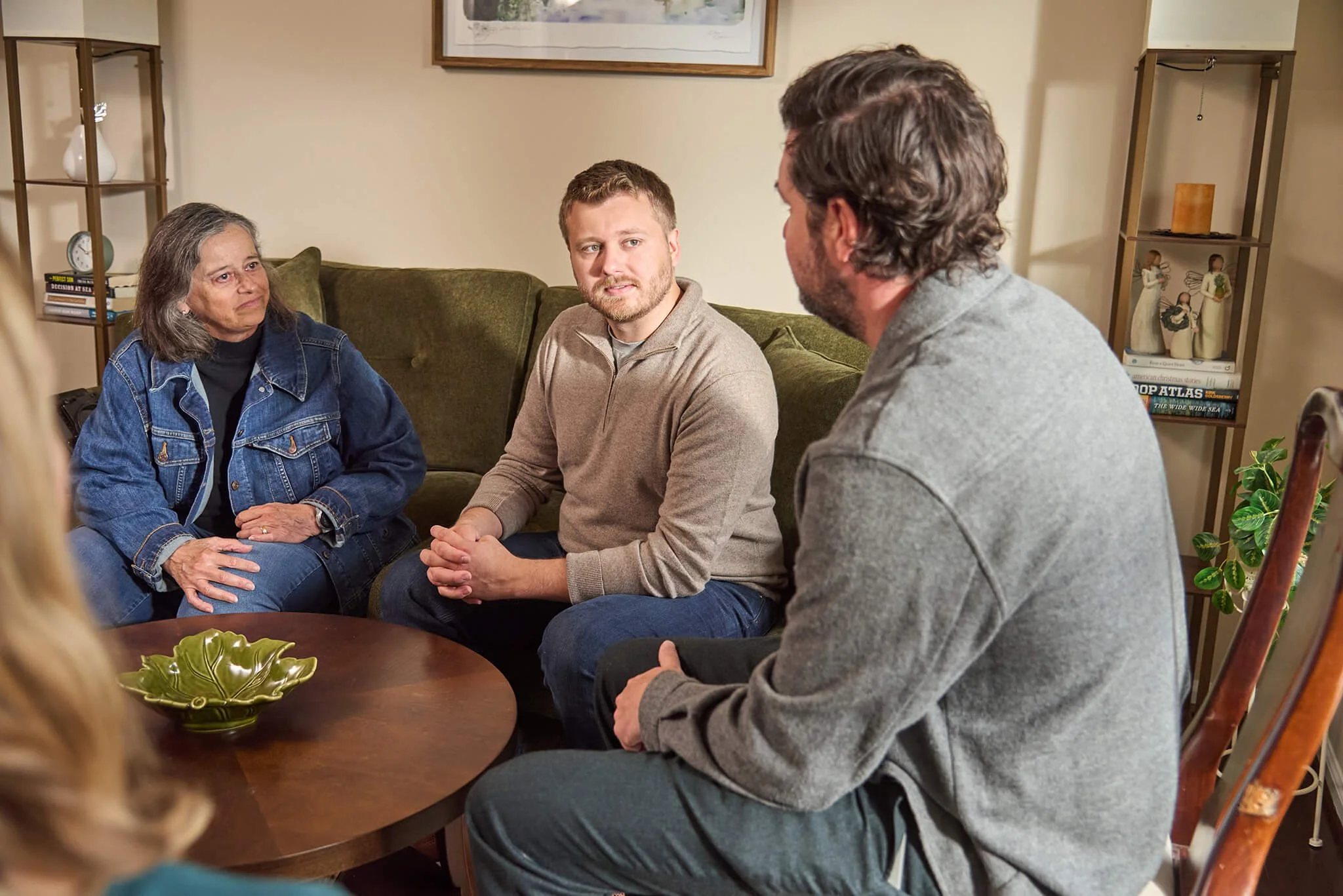  Chris walking with two people sitting on a green sofa with a wooden coffee table in front of them.