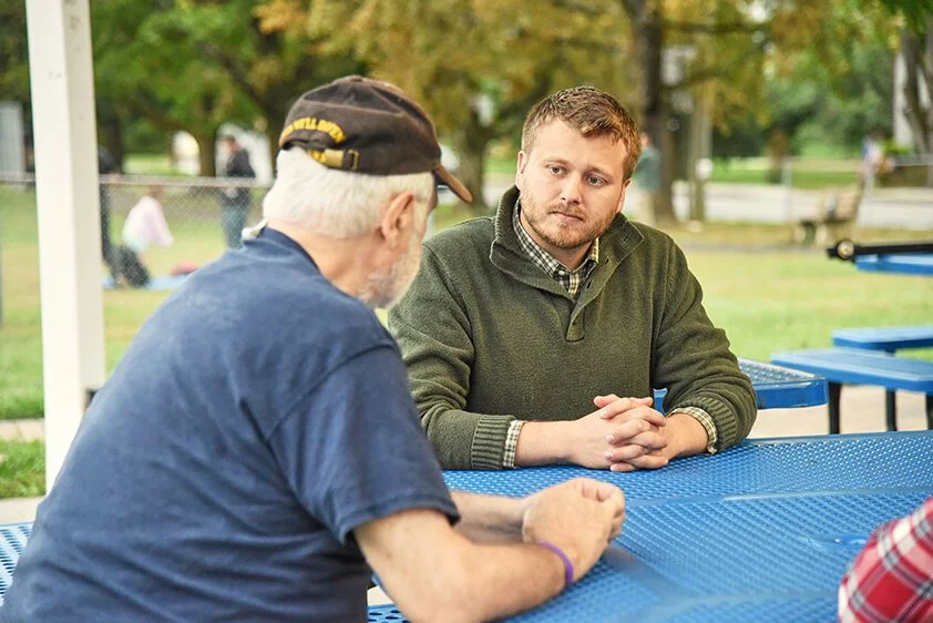 Chris talking toi a person at a blue outdoor picnic table, in a park with green trees in the background.
