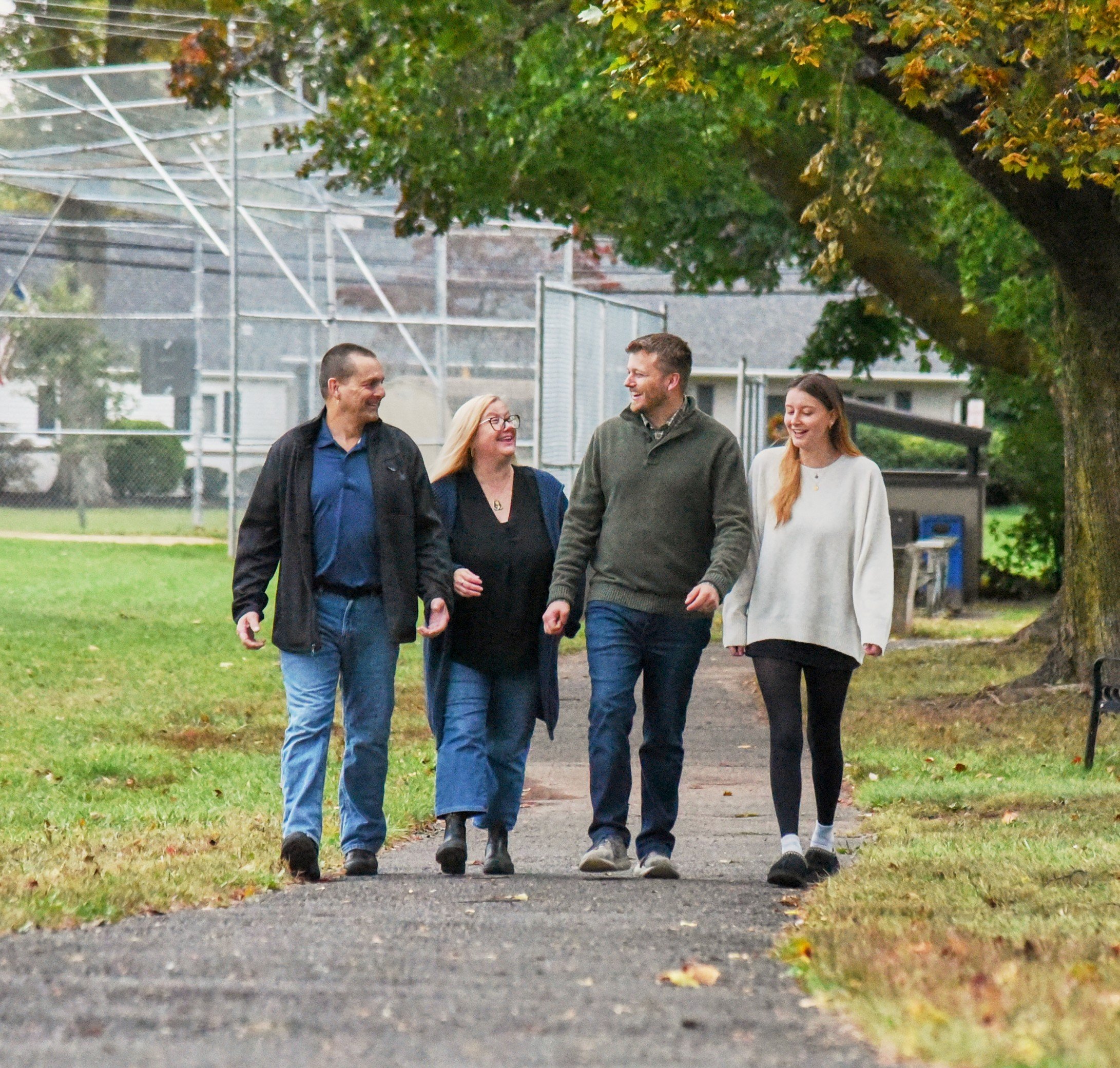 Chris walking and talking with three people on a park pathway with trees and a chain-link fence in the background.