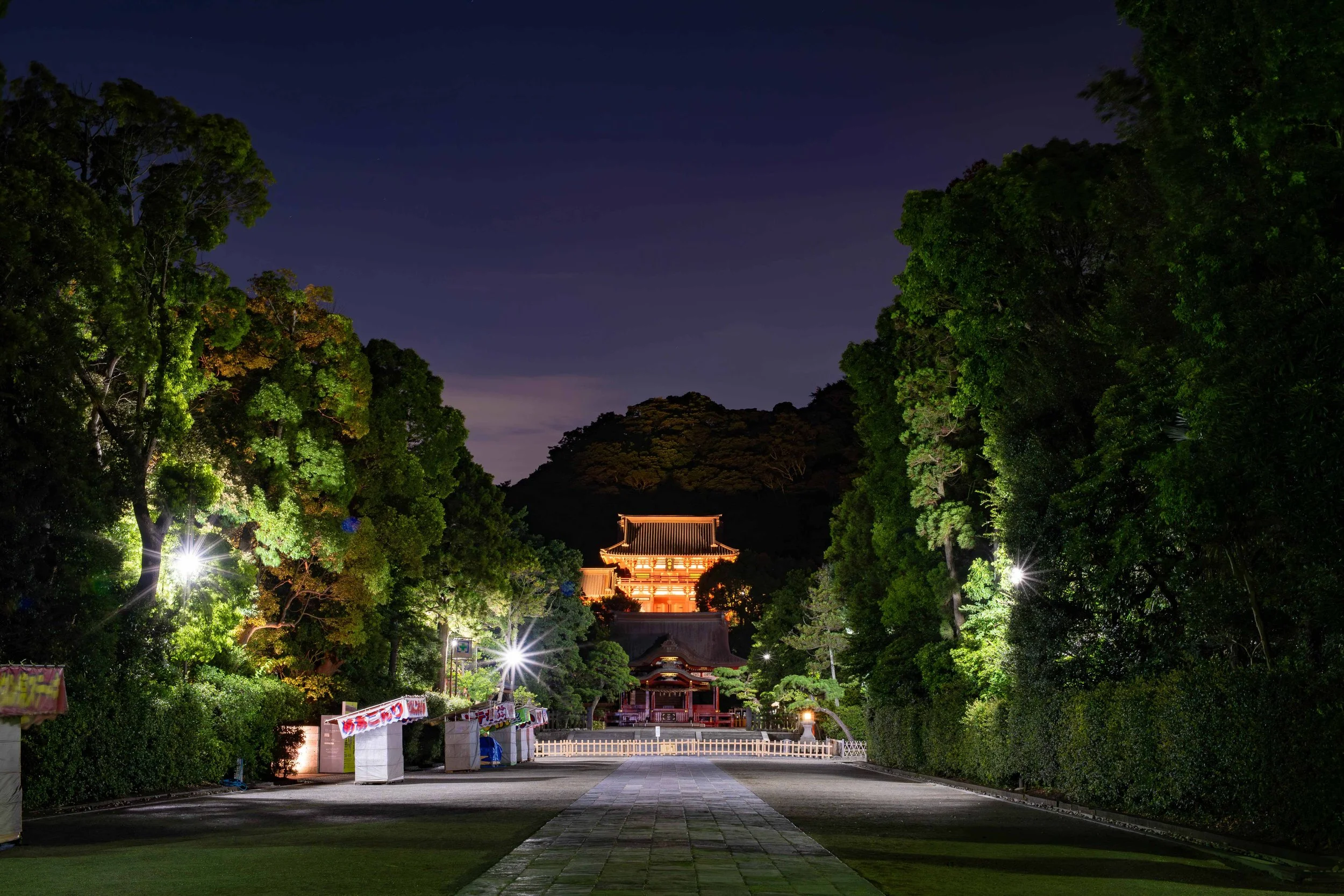 夜の神社の参道、明かりに照らされた境内と伝統的な建物が遠くに見える静かな風景