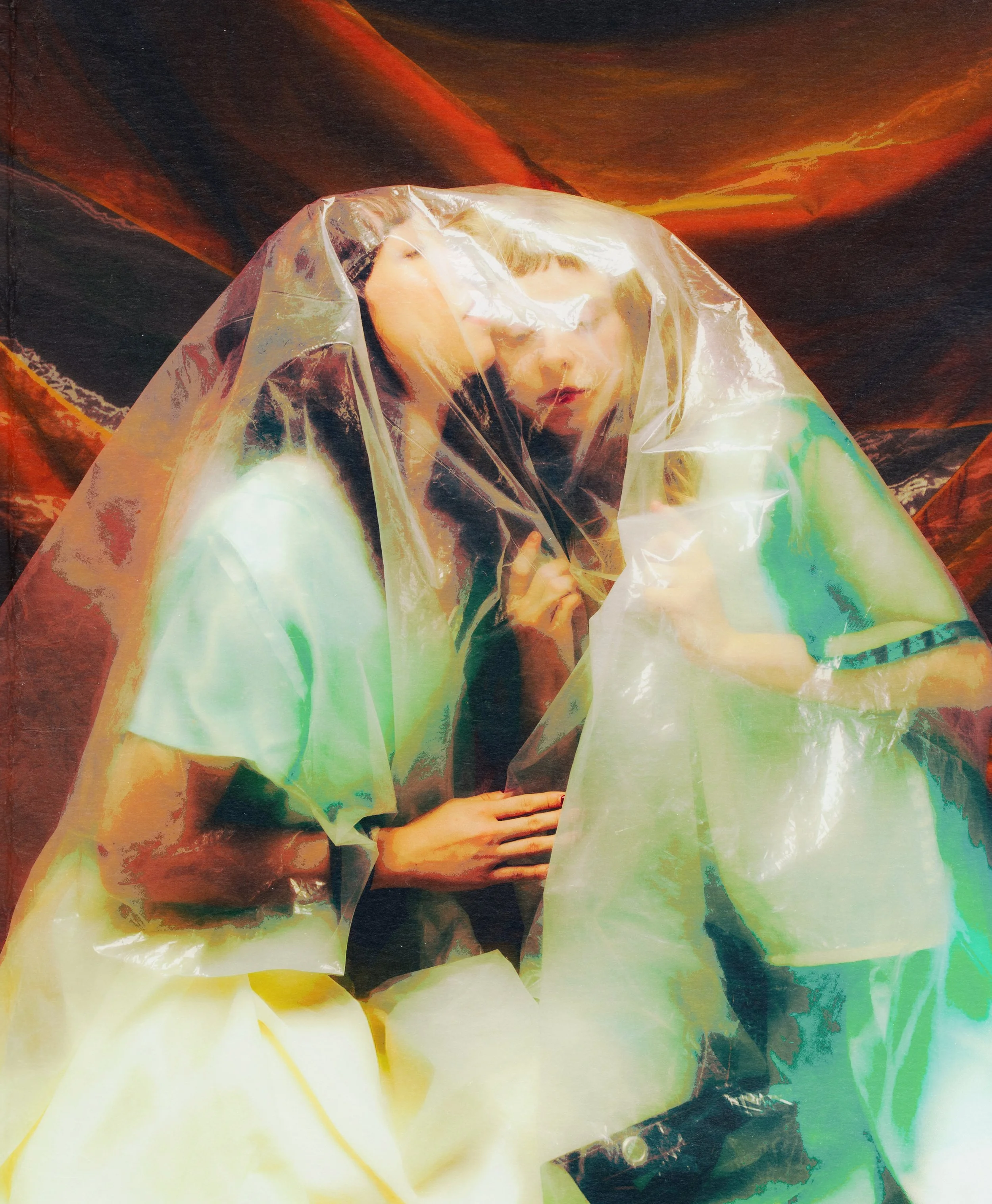Two performers sit clad in pastels under a plastic tarp, embracing.