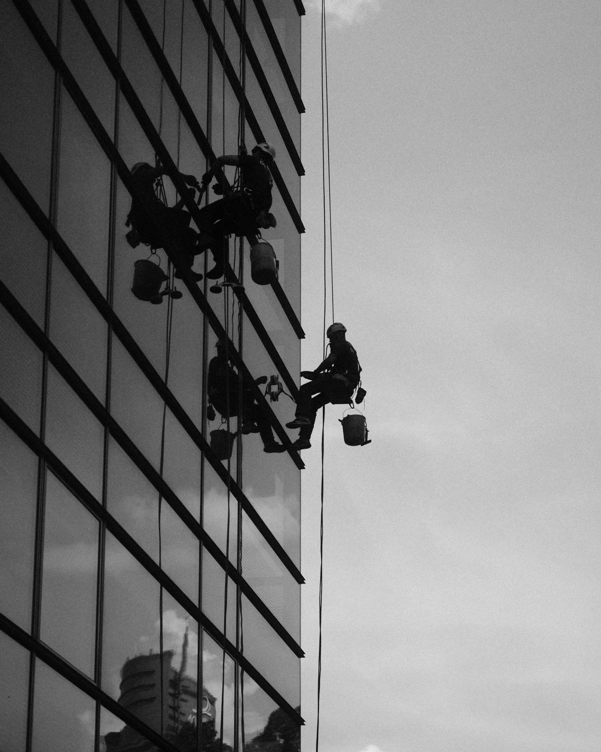 Two workers are suspended on ropes cleaning or maintaining the glass windows of a tall building. They are wearing safety helmets and harnesses, with tools and buckets attached to their harnesses.