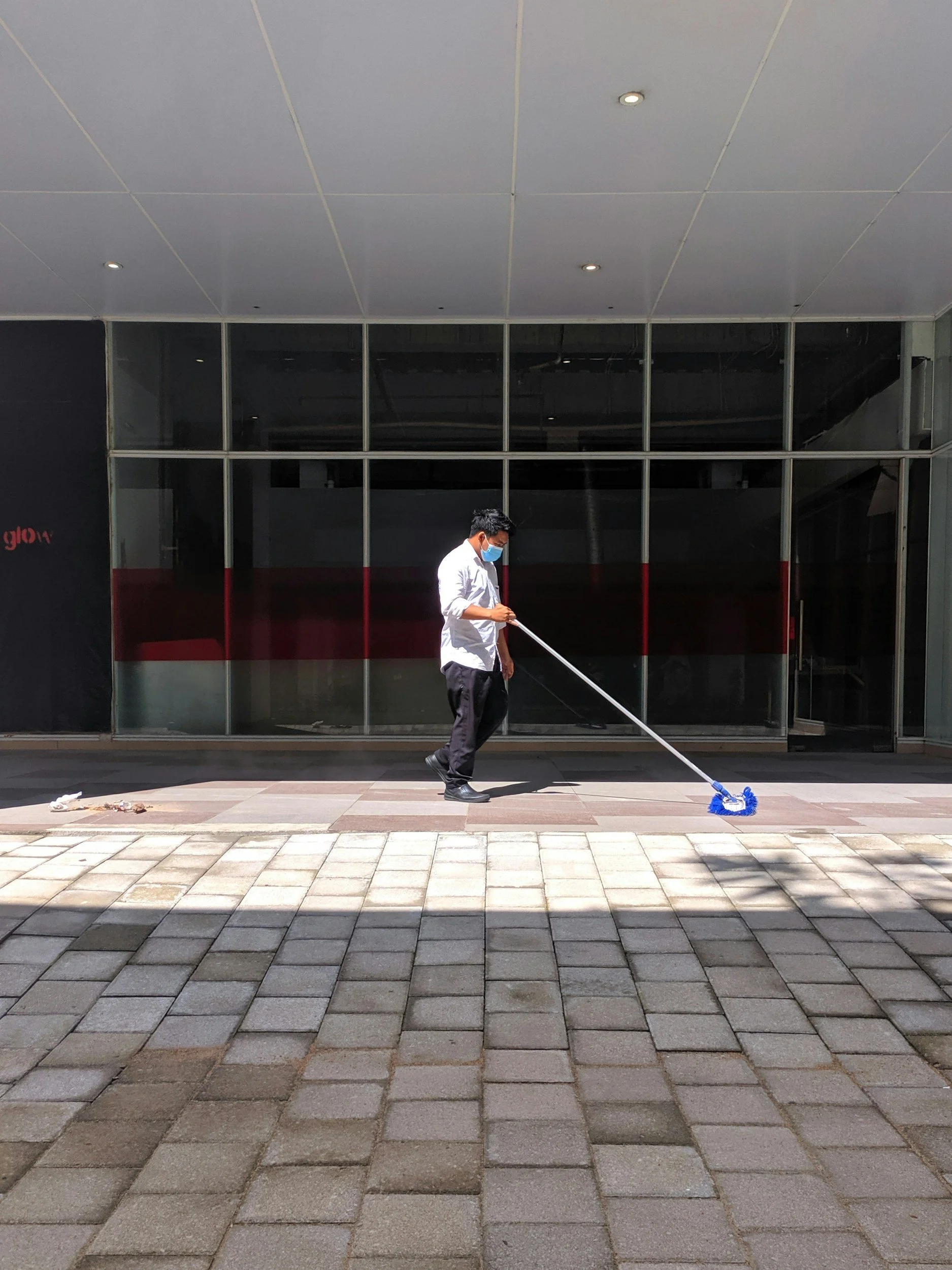 A man in a white shirt and black pants, wearing a face mask, is mopping the sidewalk outside a modern building with large glass windows.