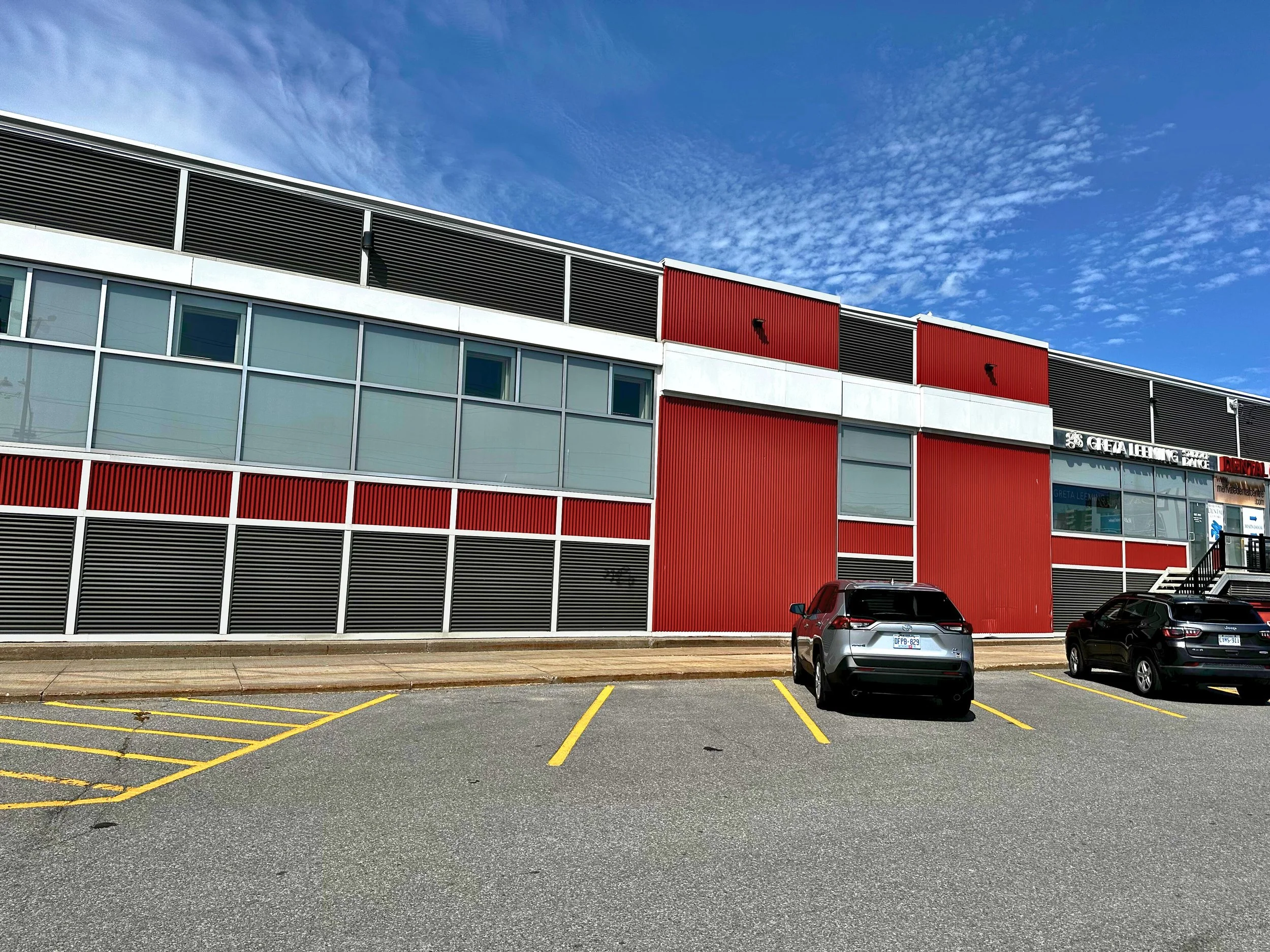 A modern building with large glass windows and red and black paneling, located in a parking lot with a few cars parked, under a partly cloudy blue sky.