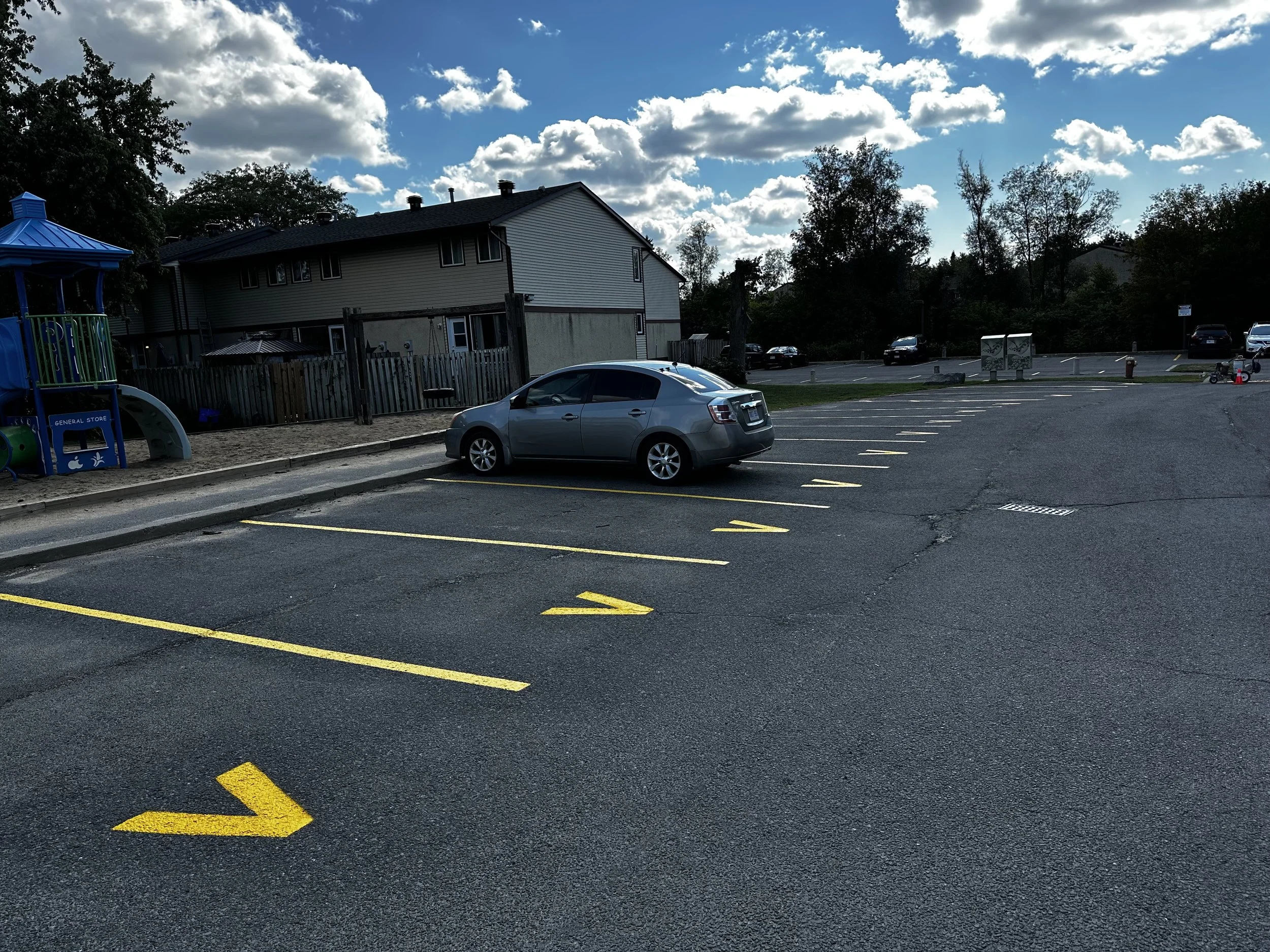 A mostly empty parking lot with one gray car parked in a space. There are bright yellow painted arrows on the asphalt indicating directions for traffic. In the background, there is a house with a gray roof, a blue playground structure on the left, trees, and some other parked cars. The sky is partly cloudy with scattered clouds.