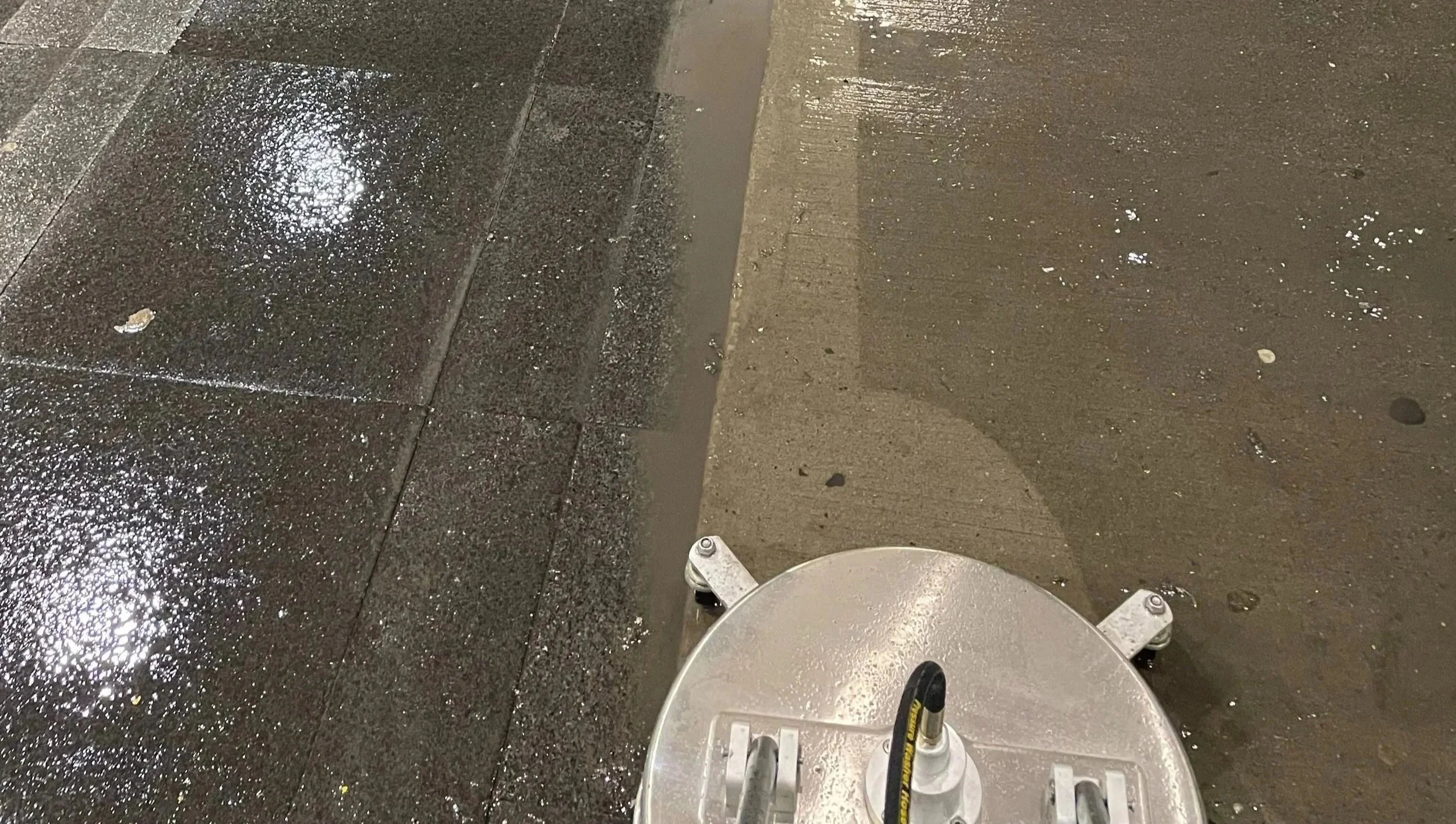 Wet sidewalk and street at night with a street cleaning machine in the foreground.