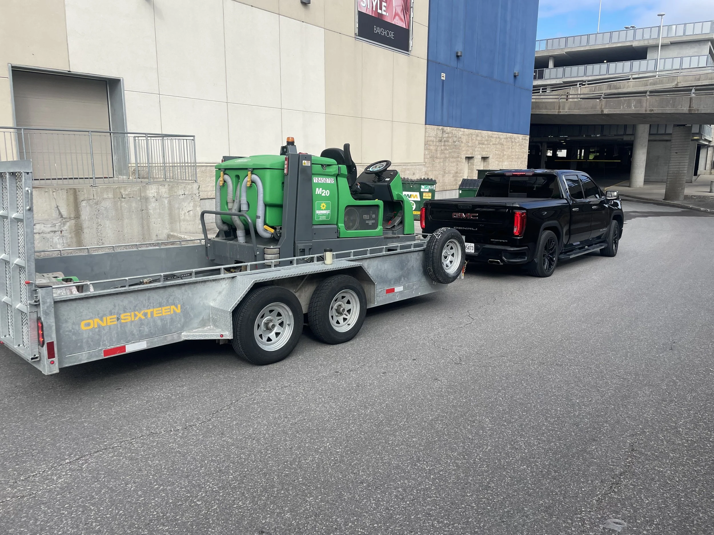 A black GMC pickup truck attached to a utility trailer carrying a green and gray piece of equipment, parked outside a modern building with blue and beige walls.