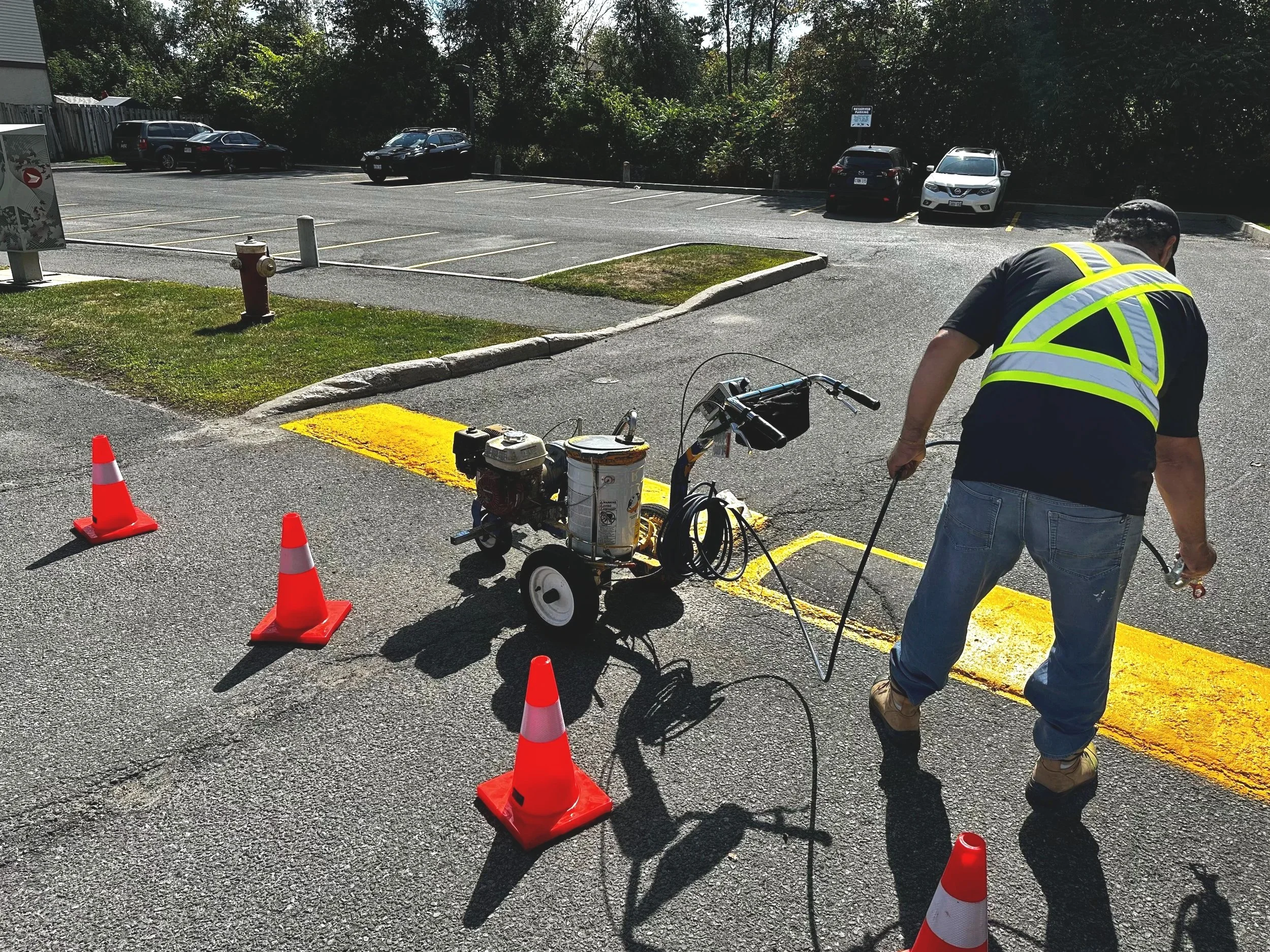Worker in a black shirt and jeans applying yellow line striping to a parking lot with traffic cones and equipment nearby.