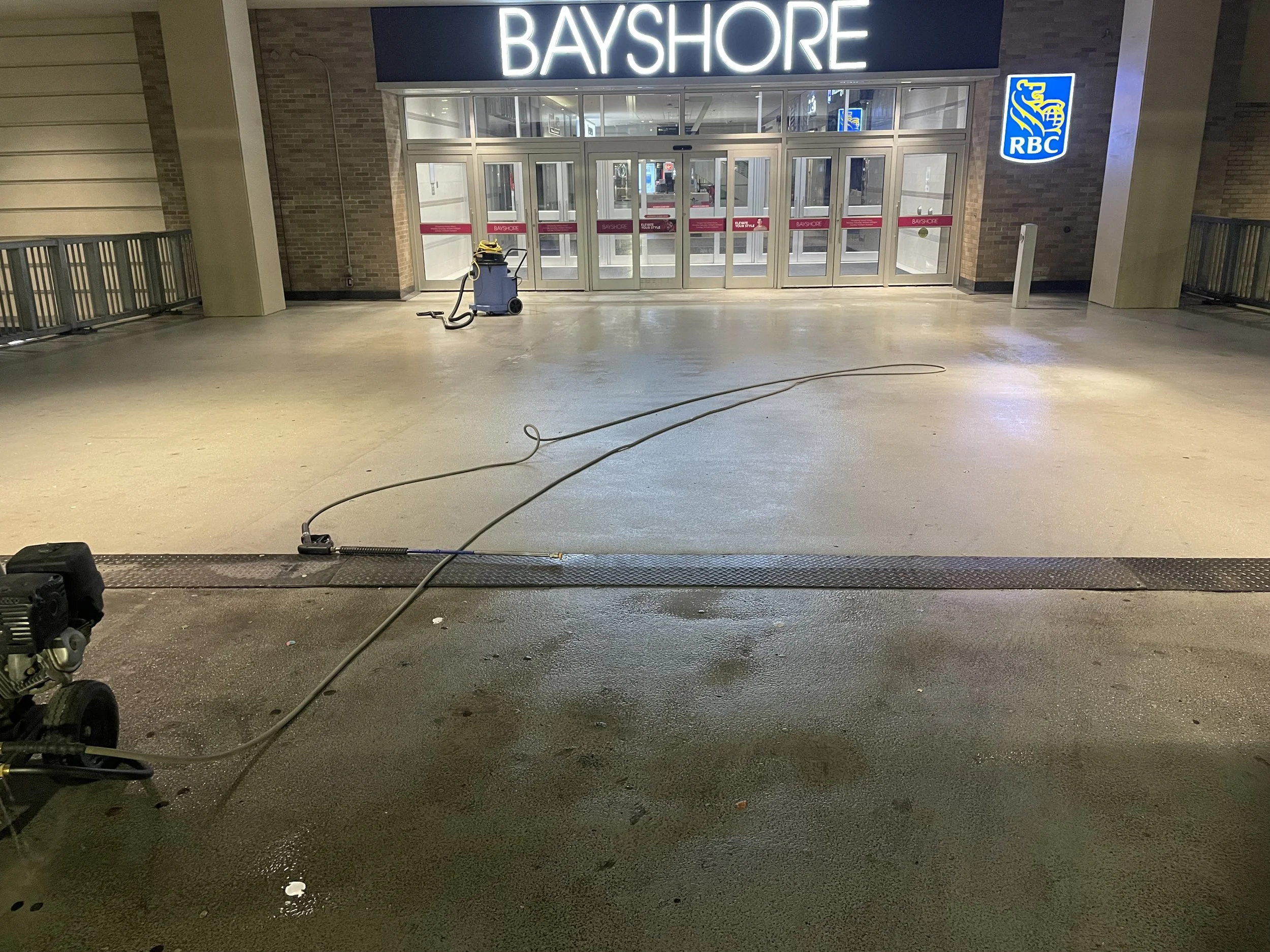 The entrance of a shopping mall or store called Bayshore, with a large illuminated sign, glass doors, and an RBC bank logo on the right side. There is a cleaning machine with a hose and water on the wet floor in front of the entrance.