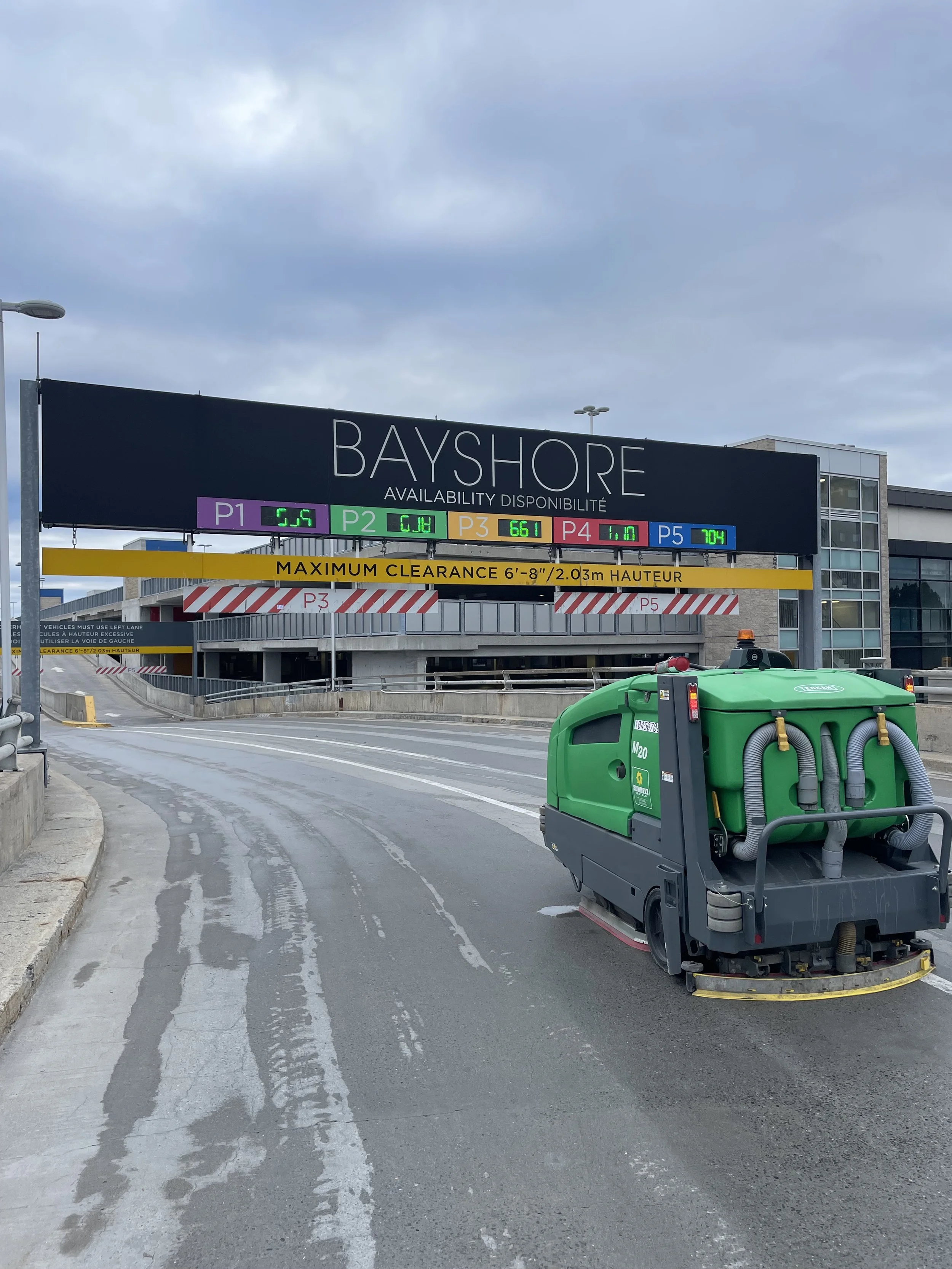 A parking garage entrance at Bayshore with an electronic display showing availability for different parking levels and a street cleaning machine in the foreground.