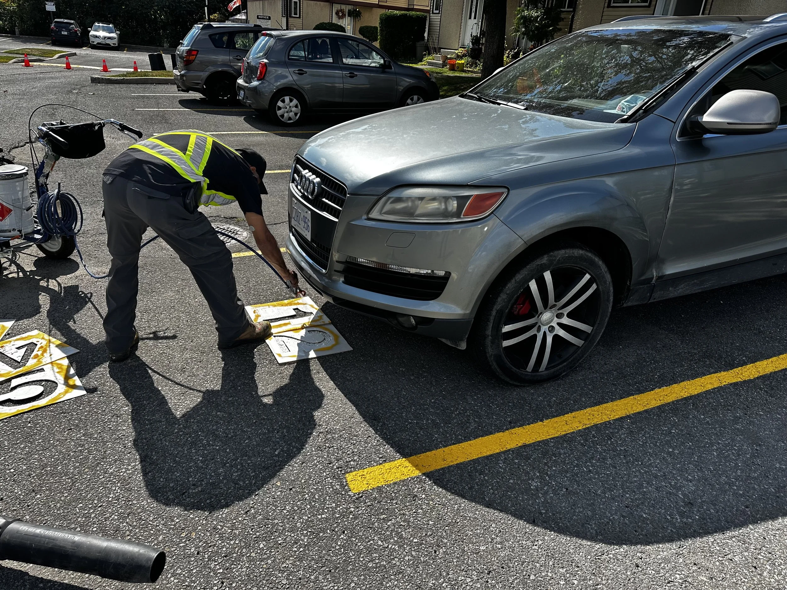 A worker in a yellow safety vest and gray pants is inspecting the front of a silver SUV in a parking lot. The worker is surrounded by numbered signs and equipment, with several parked cars and residential buildings in the background.