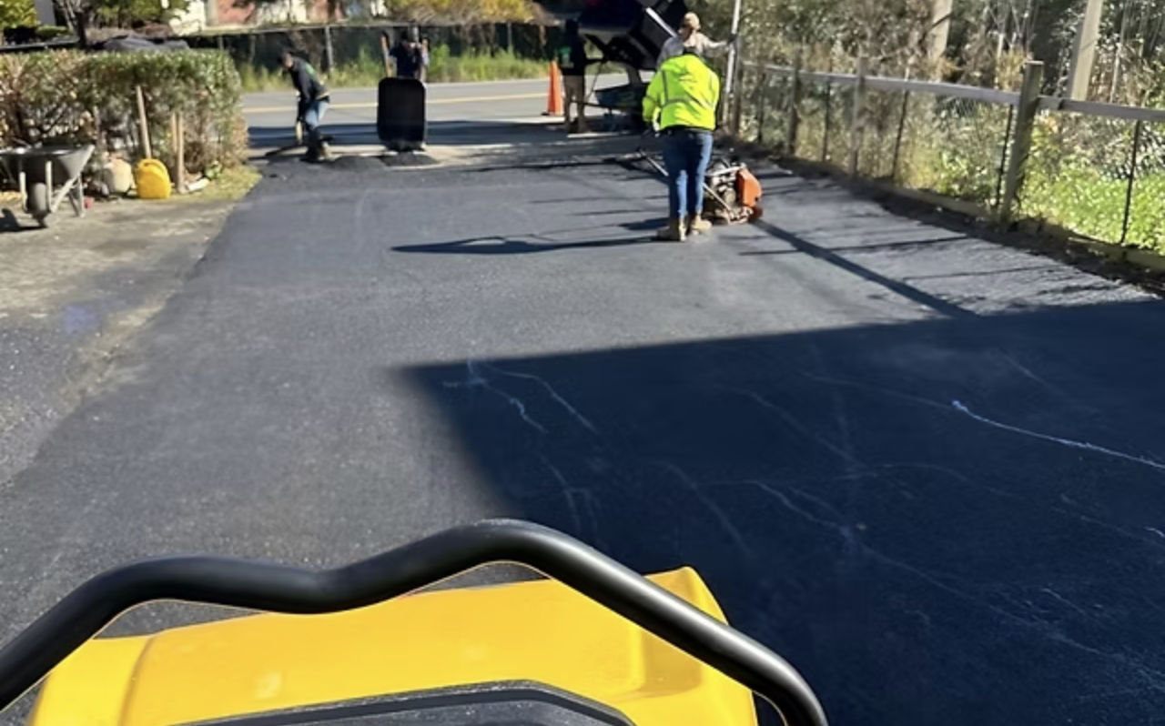Construction workers are resurfacing a road with asphalt, using machinery and tools. There is a person in a yellow safety vest operating equipment, and other workers nearby.