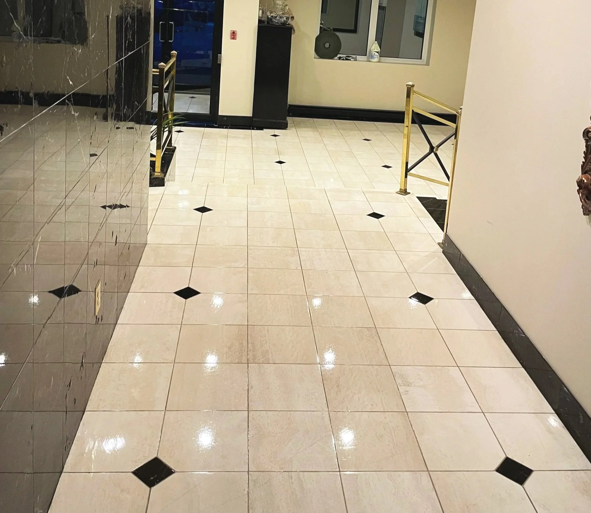 Empty hotel lobby with beige tiled floor and black diamond accents, gold-colored railing on the right, black and beige walls, and a glass door at the back.