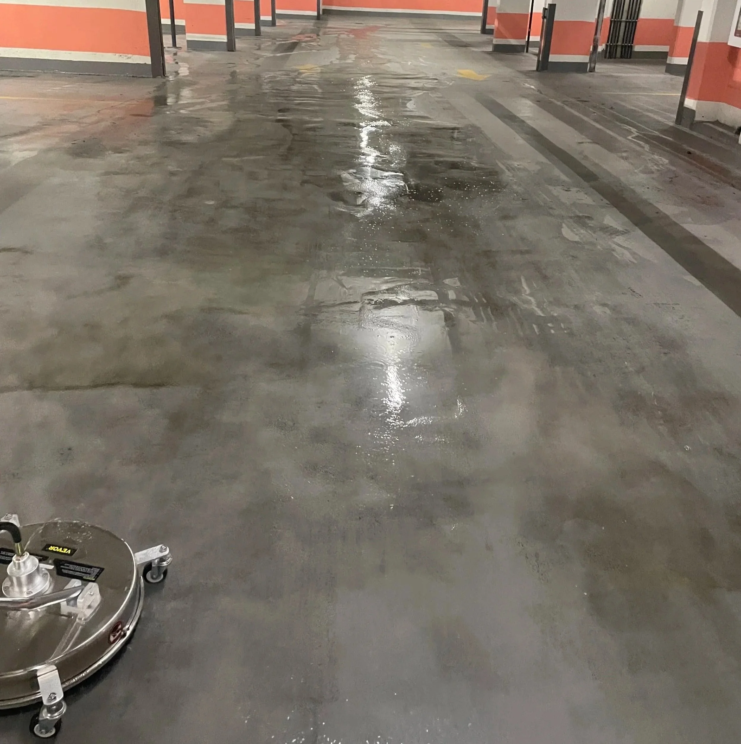 An indoor parking garage floor being cleaned with a floor cleaning machine, with water and cleaning solution visible on the concrete surface.