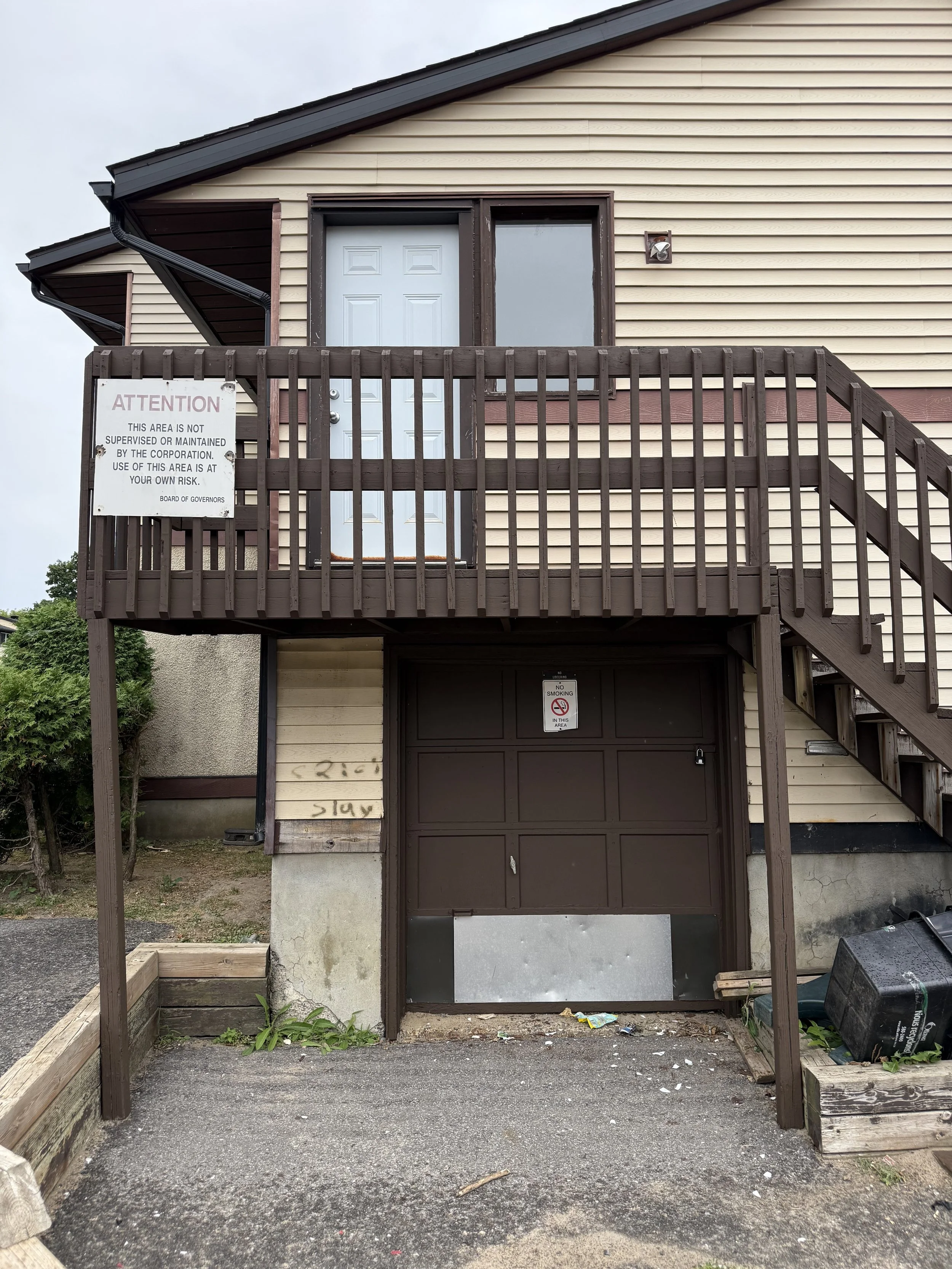 A two-story house with beige siding and dark brown trim. There is an elevated deck with a railing and a door leading into the house, and a staircase on the right side. The garage door on the ground level is dark brown, with a sign that says 'No Smoking.' There is a sign on the deck railing that warns of the area not being supervised. The driveway has some debris and construction materials nearby.