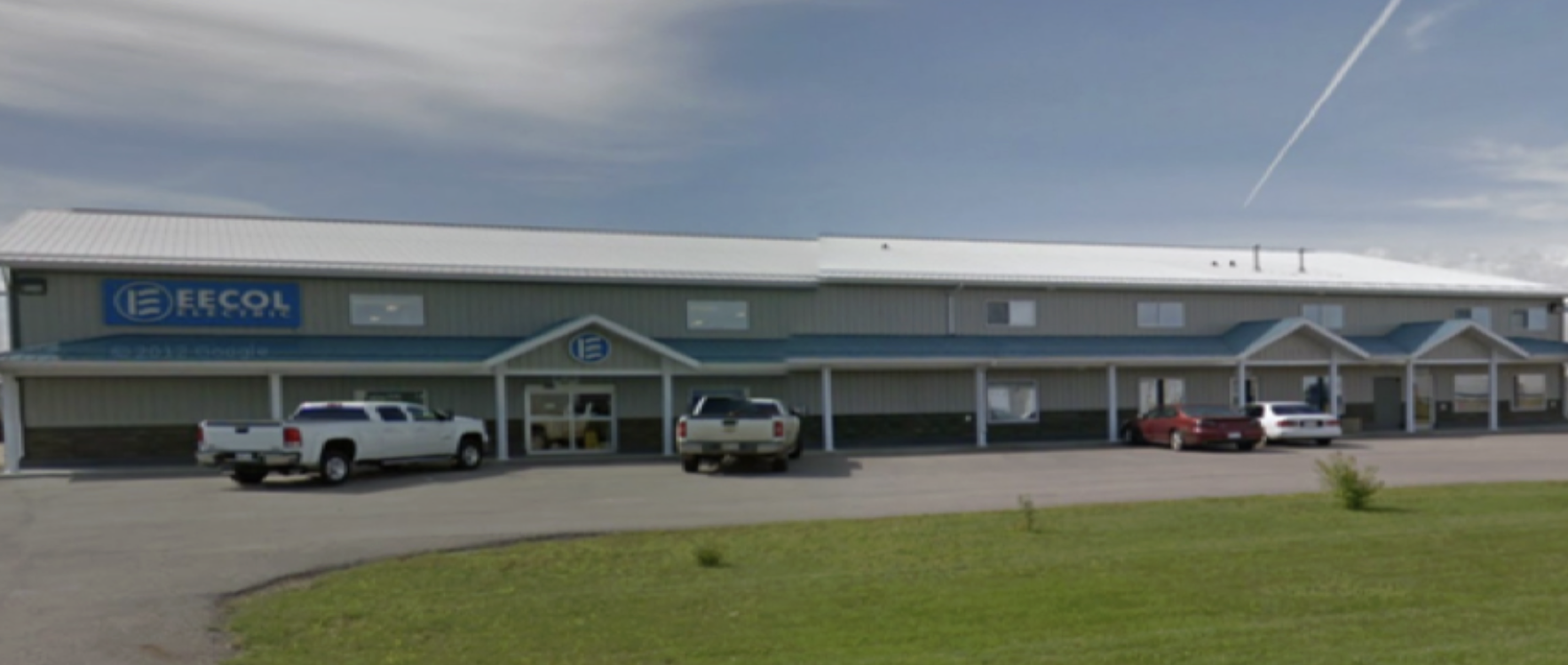 An industrial building with a blue sign that says 'EECOL electric,' parked cars, and a grassy area in the foreground.