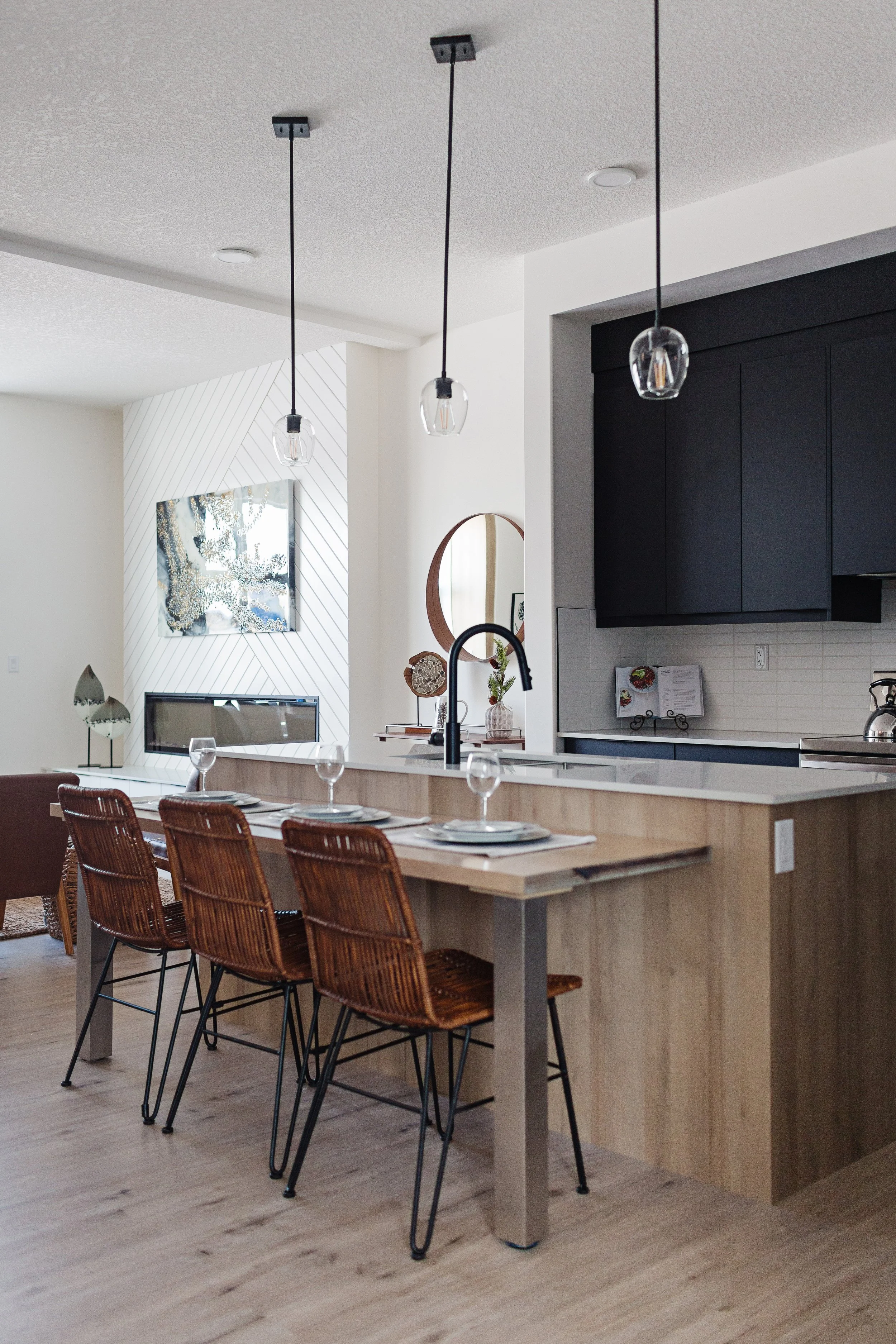 Modern open-concept kitchen and living room with a wooden bar counter, four rattan bar stools, black overhead pendant lights, and a white wall with decorative art and a mirror.