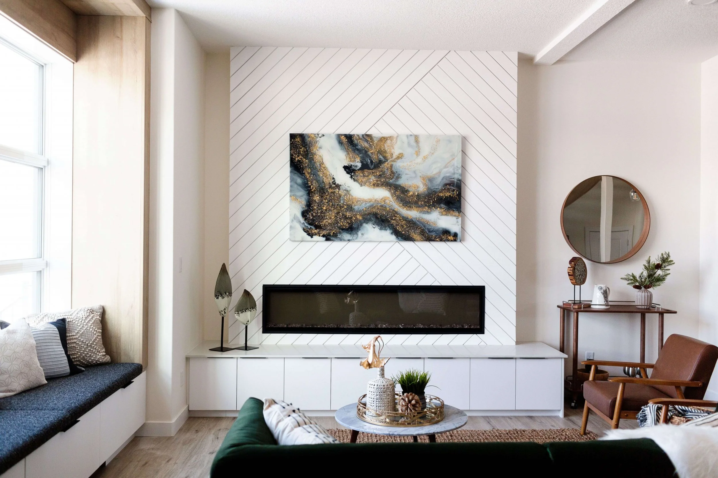 Living room with a white accent wall featuring diagonally lined paneling, a rectangular fireplace, a colorful abstract painting, a circular mirror, wooden furniture, and decorative plants.
