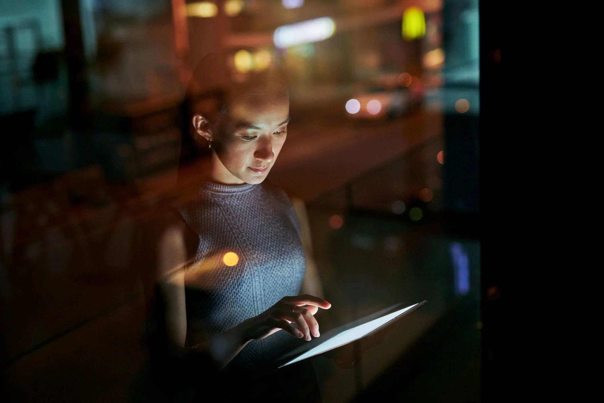 A woman using a tablet at night, seen through a window with reflections of city lights and traffic.