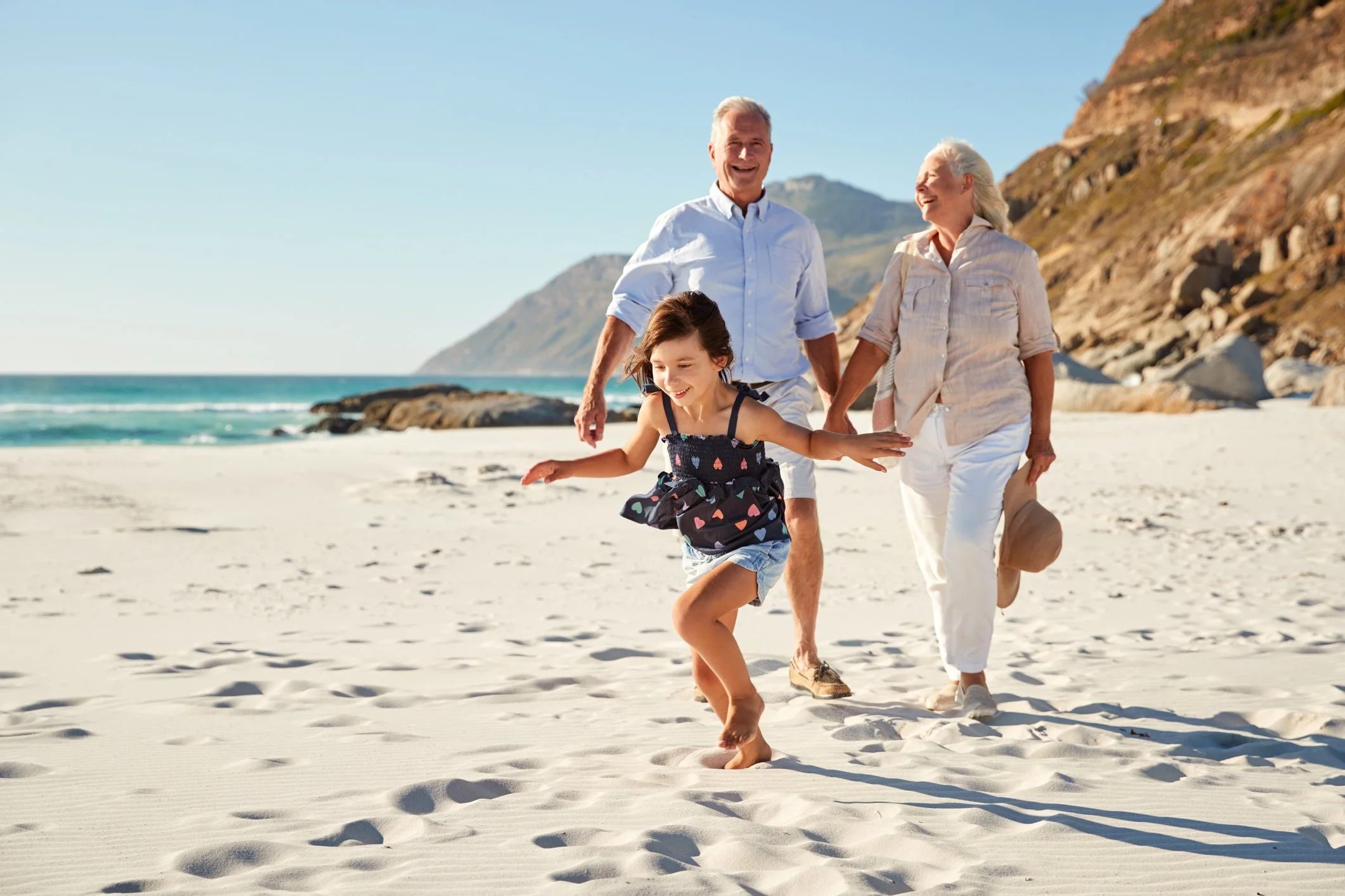 A family of three enjoying a day at the beach, with a young girl running on the sand and her grandparents walking behind her, ocean waves and a mountain in the background.