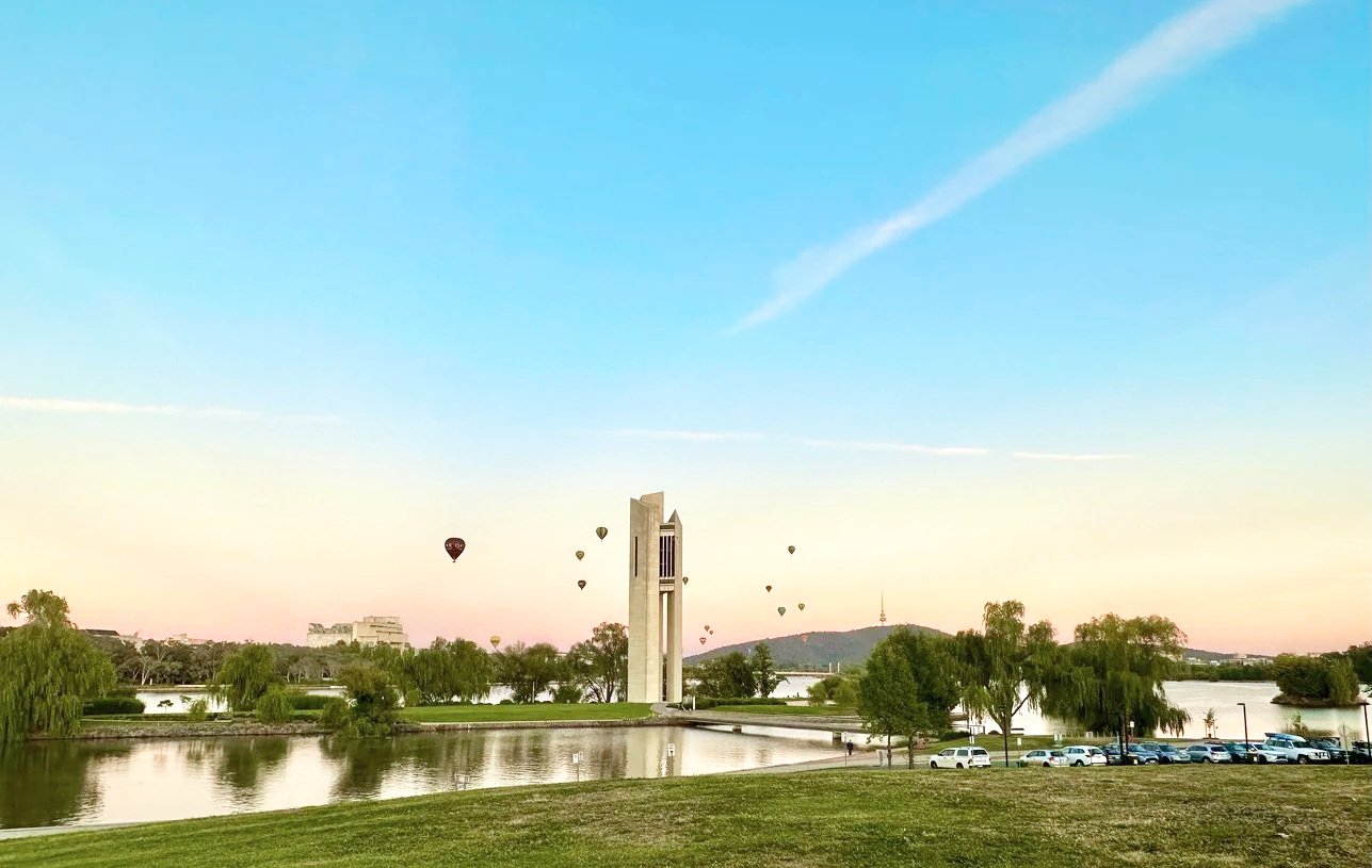 A park with a lake, trees, and a tower in the background, with multiple hot air balloons floating in the sky during sunset.