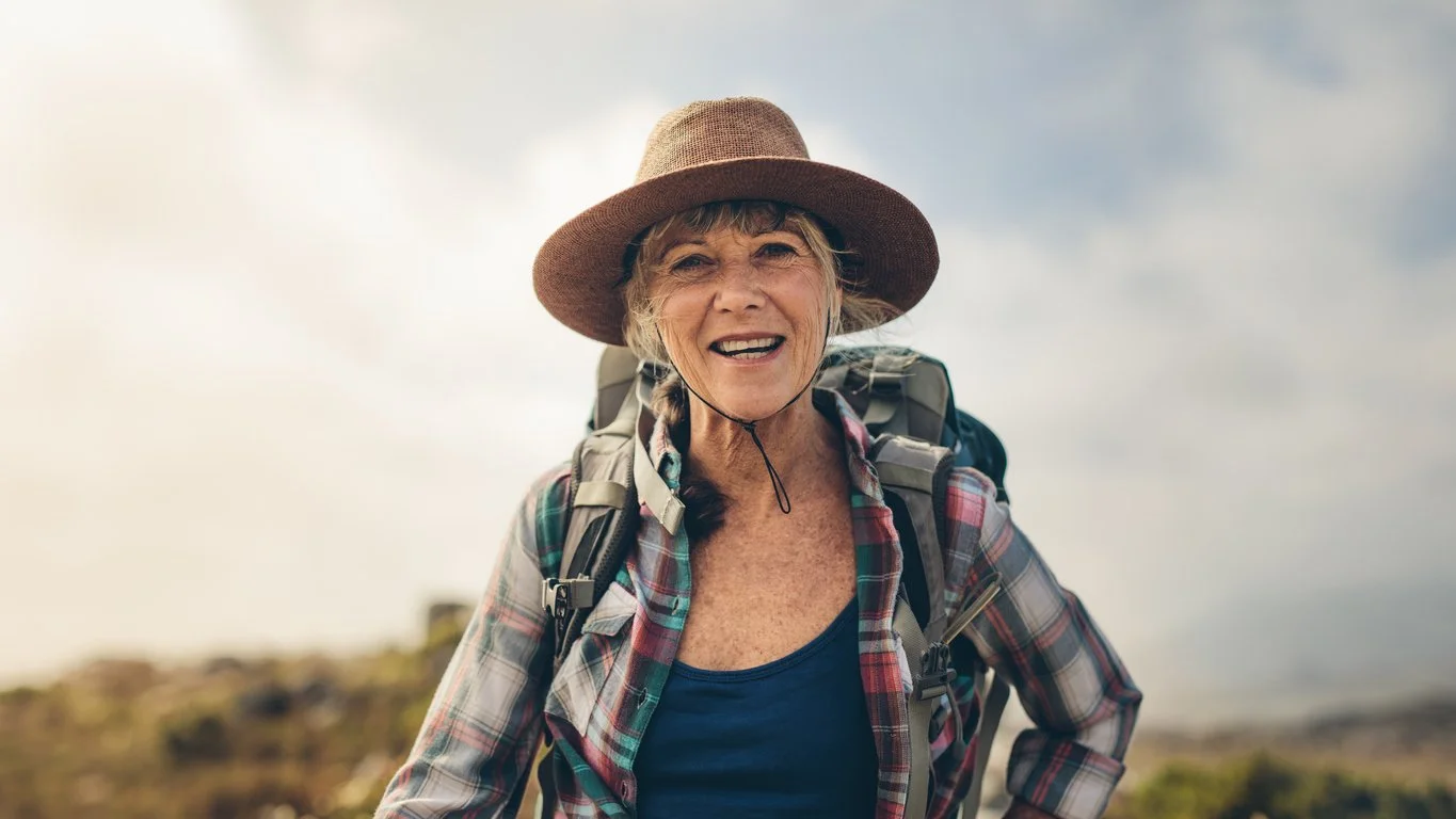 An elderly woman wearing a wide-brimmed hat, plaid shirt, and backpack outdoors, smiling at the camera.
