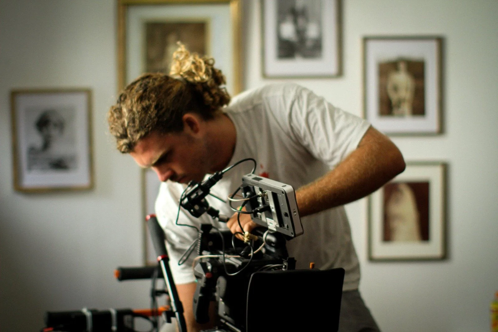 A man with curly hair working on a camera setup in a room with framed photographs on the wall.