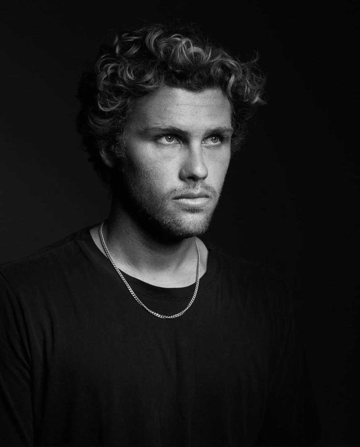 Black and white portrait of a young man with curly hair, wearing a black t-shirt and a silver chain necklace, looking to the side with a serious expression.