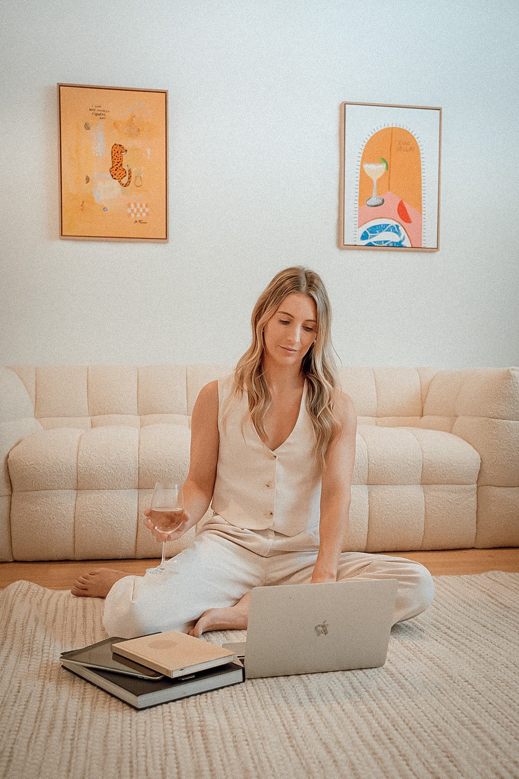 A woman in beige pajamas sitting on the floor with a laptop, holding a wine glass, with books nearby, in a living room with two framed artworks on the wall.