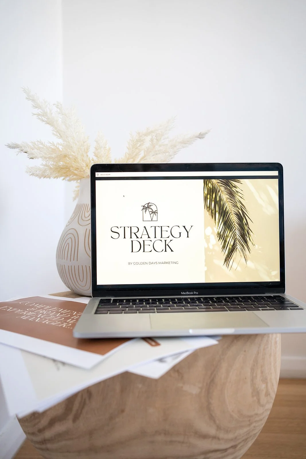 Laptop on a wooden table displaying a presentation titled 'Strategy Deck' by Golden Days Marketing, with a background image of a palm leaf. Behind the laptop is a white vase with decorative white dried plants.