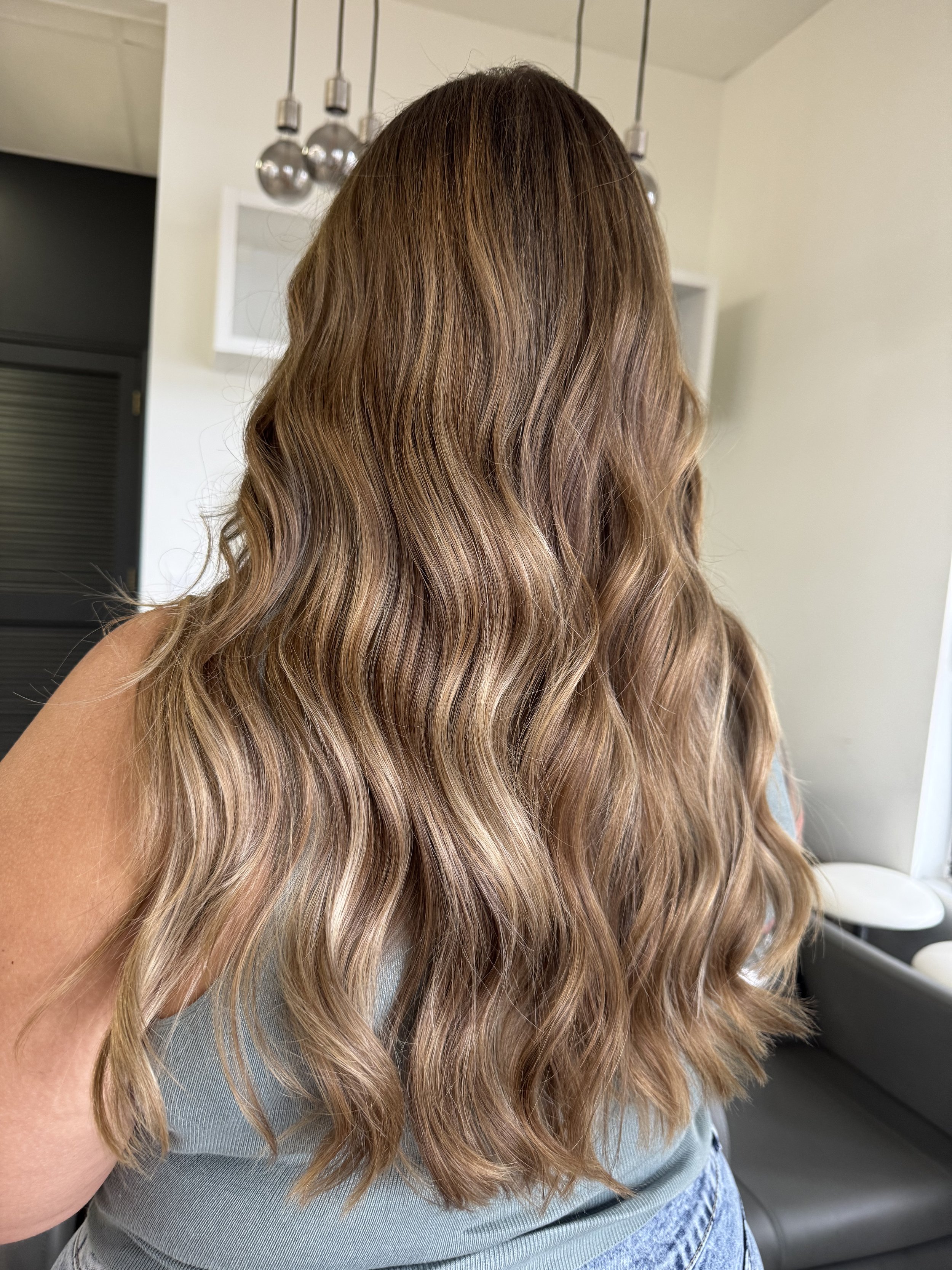 Back view of a woman with long, wavy, light brown hair standing indoors.