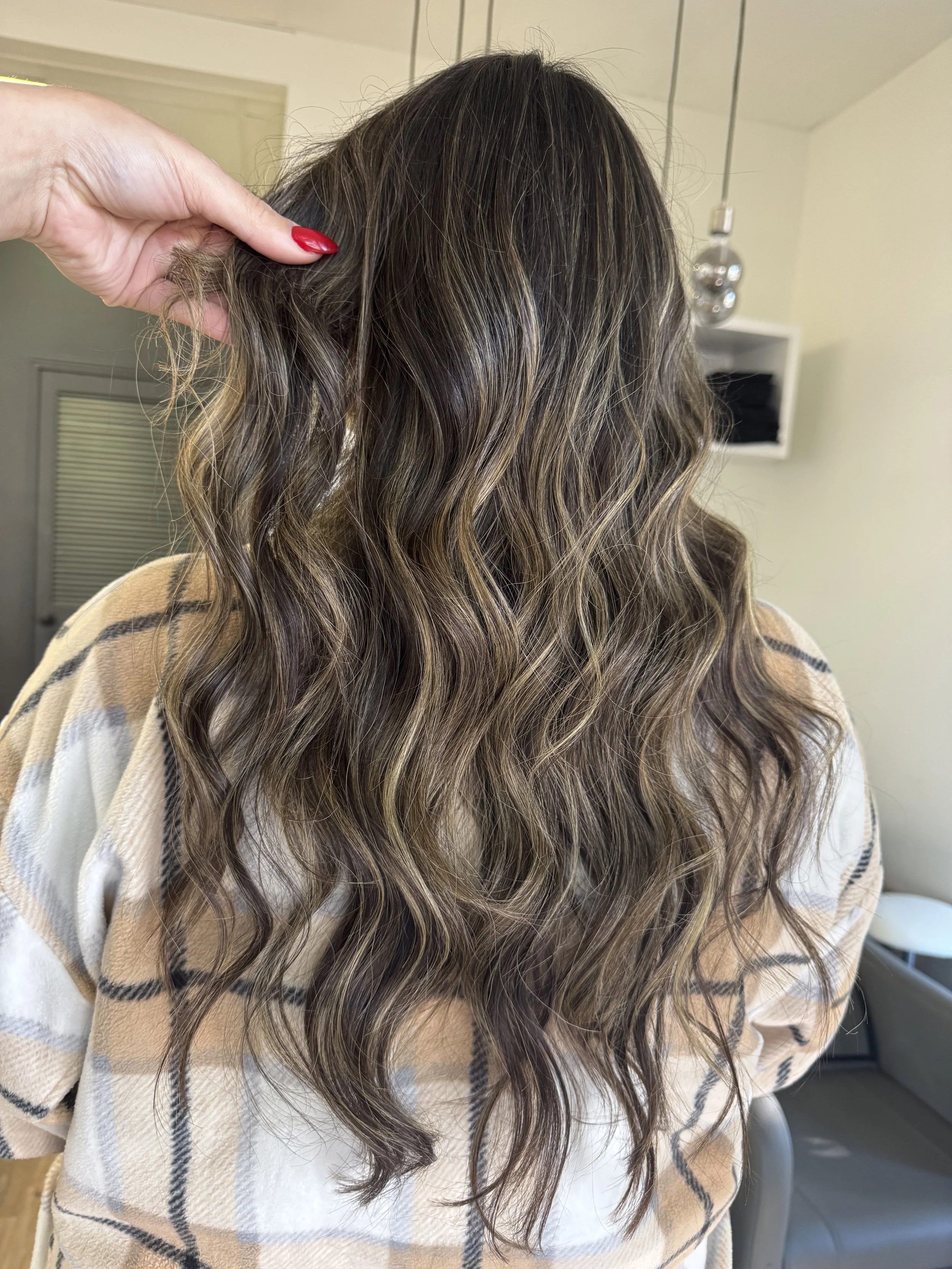 Back view of a woman with long, wavy, brown hair with blonde highlights, wearing a beige and white plaid shirt in a salon setting.