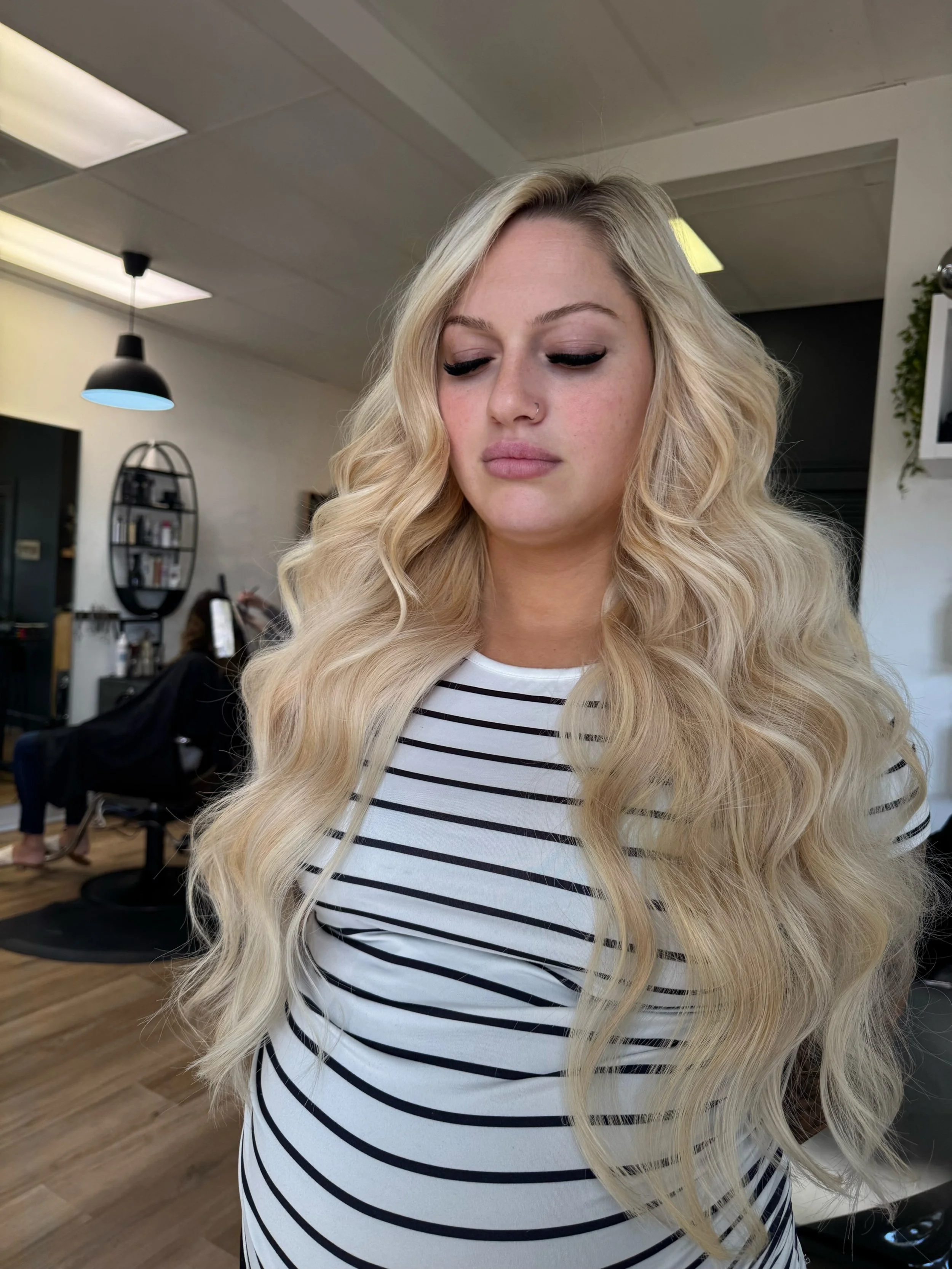A woman with long, blonde, wavy hair and light makeup, wearing a white shirt with black horizontal stripes, standing in a salon.