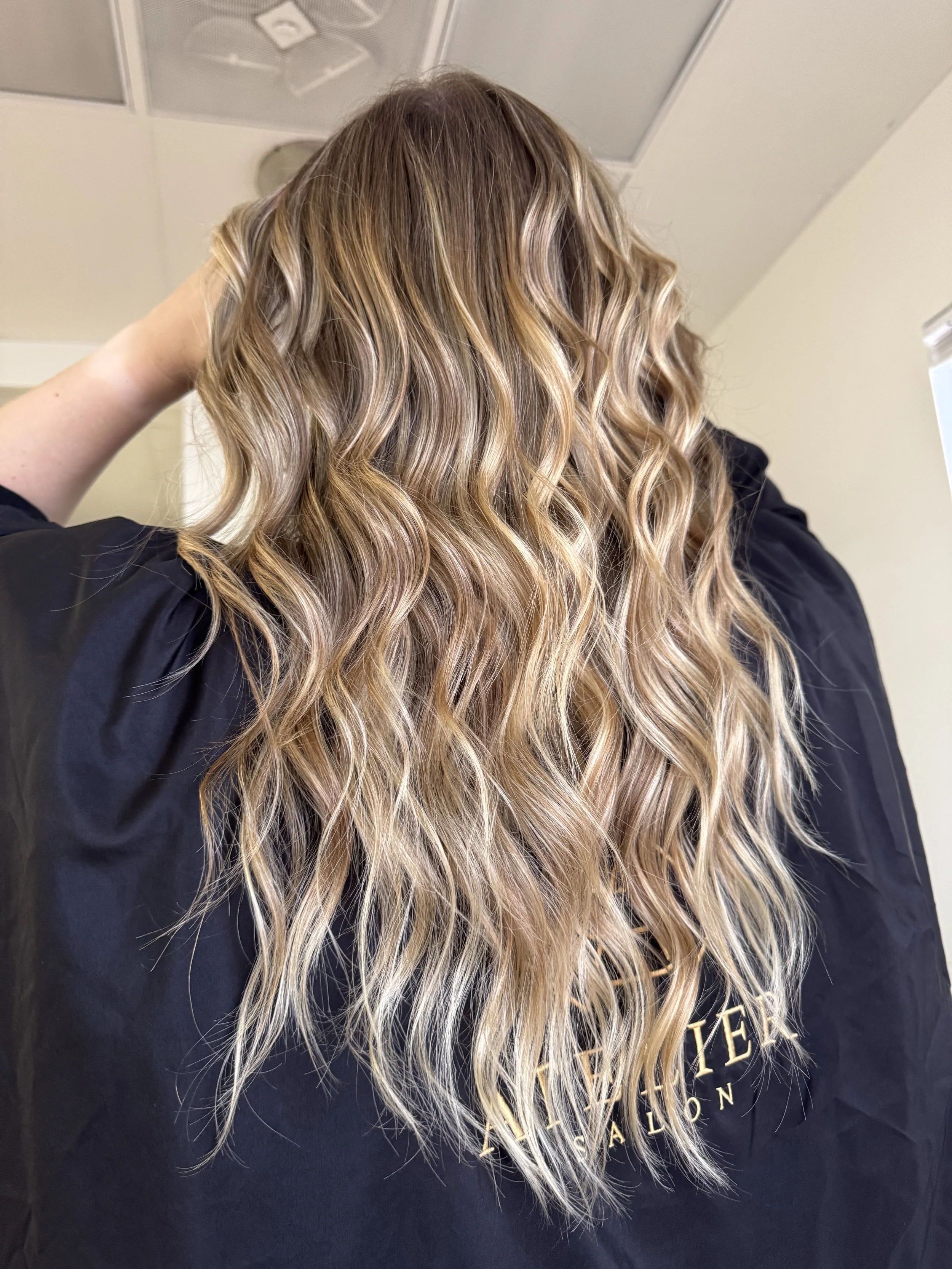 Long, wavy blonde hair with highlights seen from the back, worn by a person wearing a black cape or salon gown.