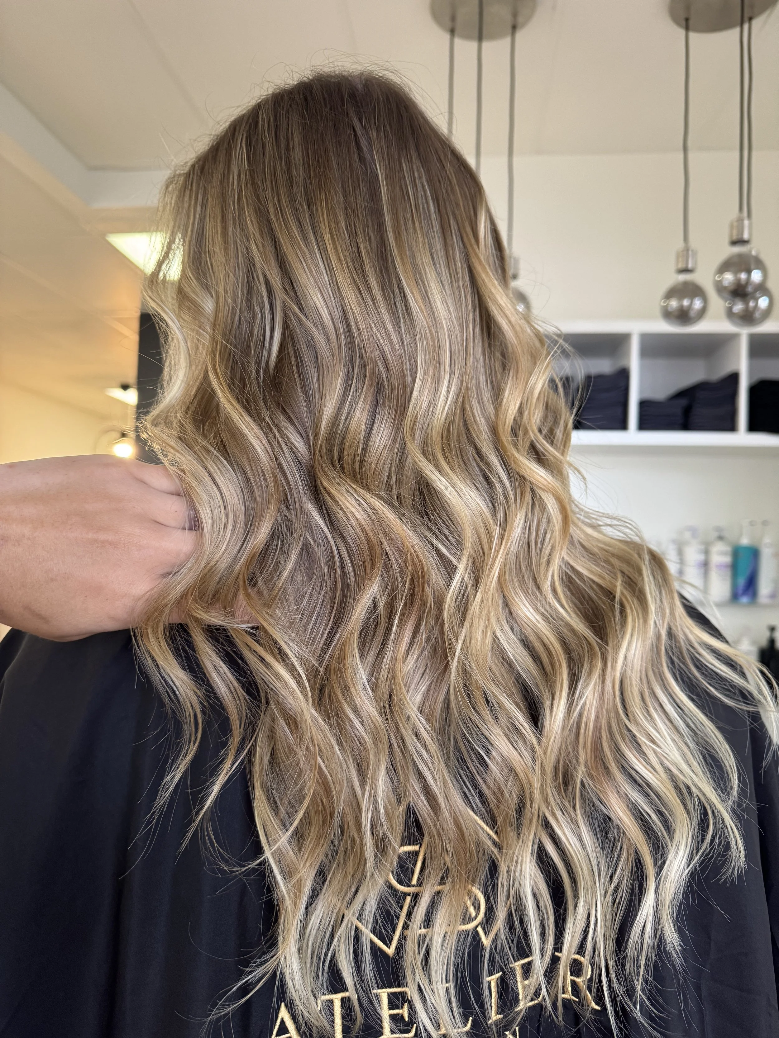 A person with long, wavy, blonde hair, seen from the back in a hair salon.