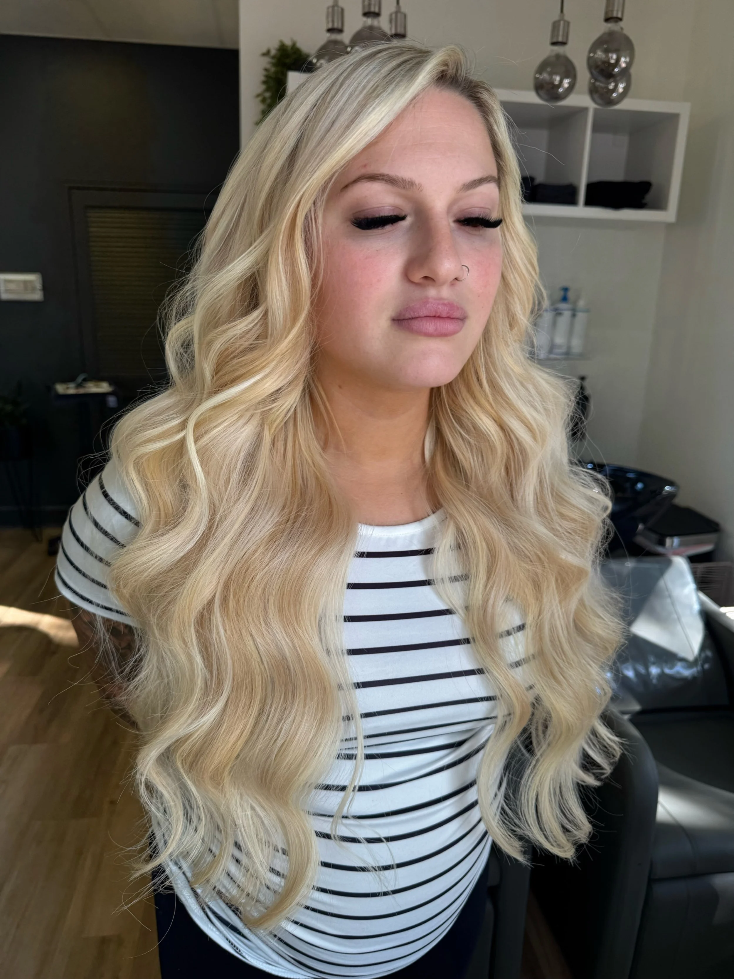 A woman with long, wavy blonde hair, wearing a white and black striped shirt, with her eyes closed, standing in a salon or studio with hair styling products and equipment behind her.