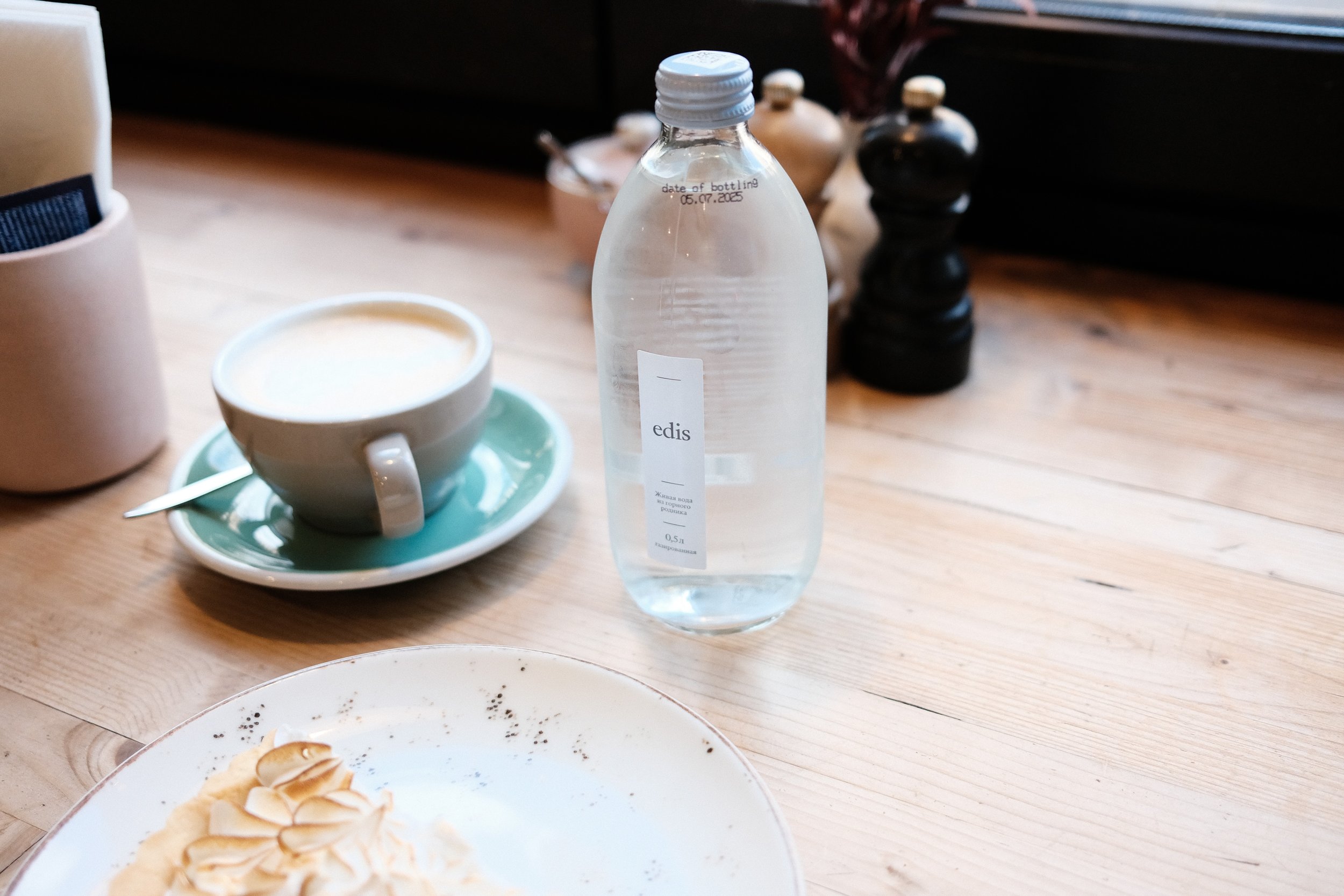 A table setting with a plate of dessert, a cup of coffee with a spoon, a bottle of water, and salt and pepper shakers.