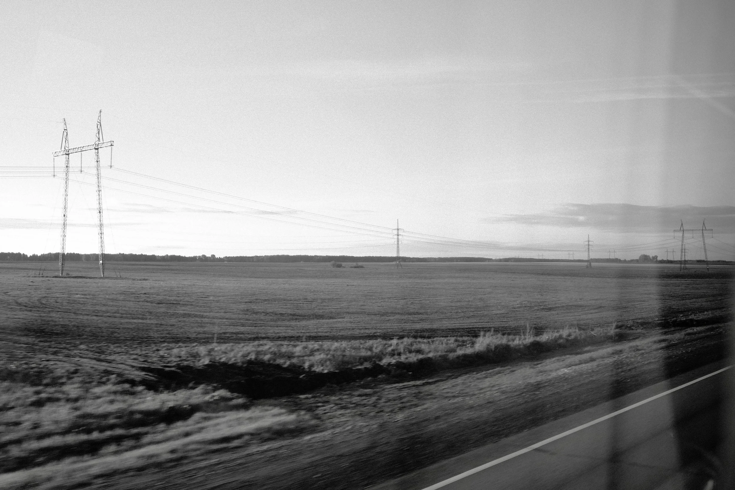 Black and white photo of a rural landscape with power lines and a clear sky, taken from a moving vehicle.