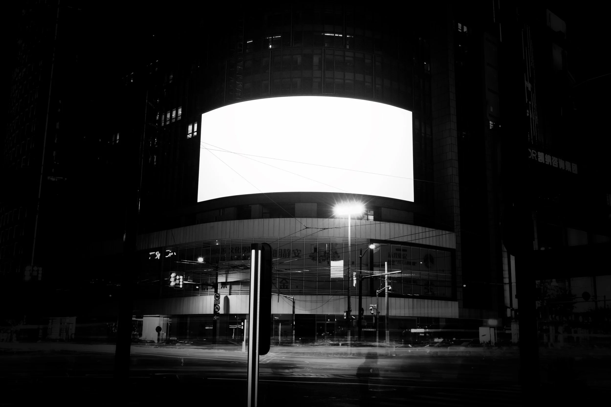 Nighttime city scene featuring a large illuminated billboard on a modern building, with streetlights and power lines visible below.