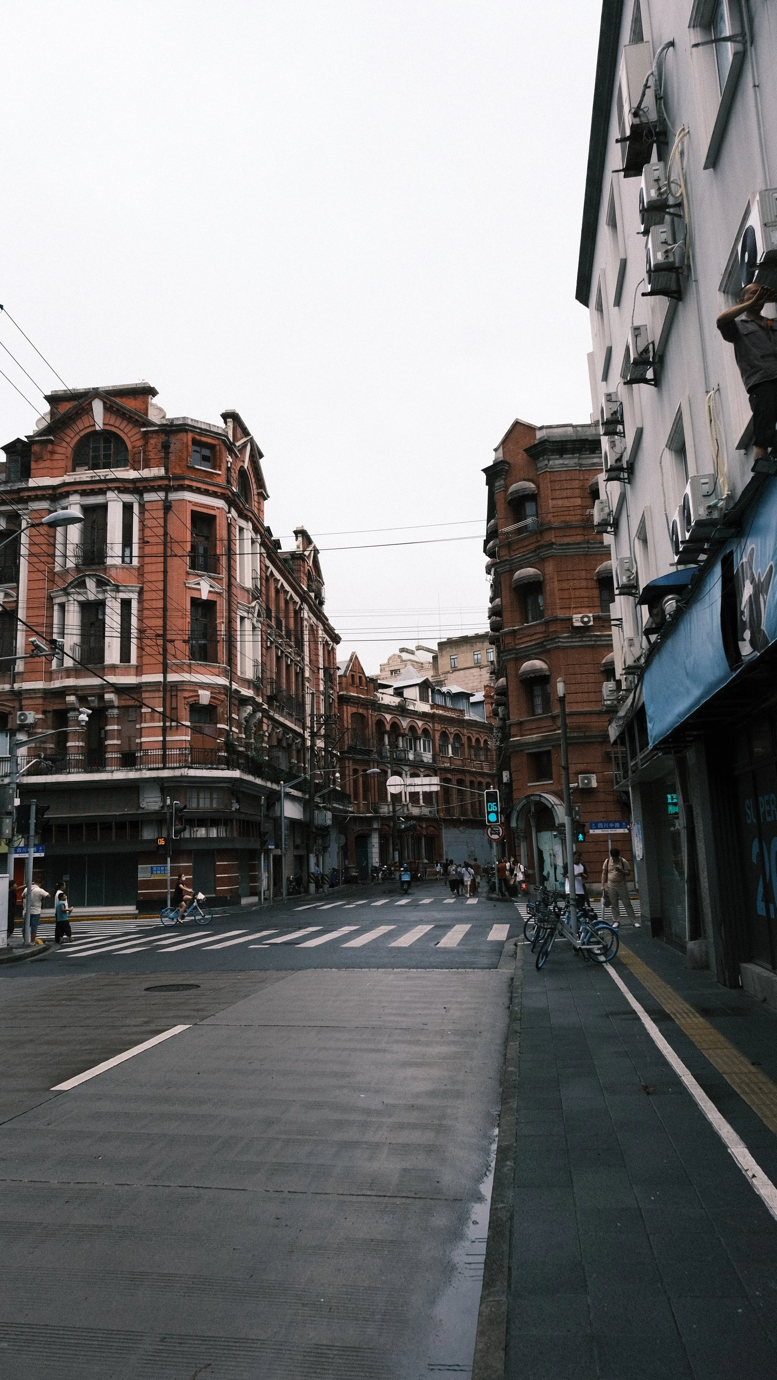 City street scene with old brick buildings, pedestrians, bicycles, and overhead electrical wires, under an overcast sky.