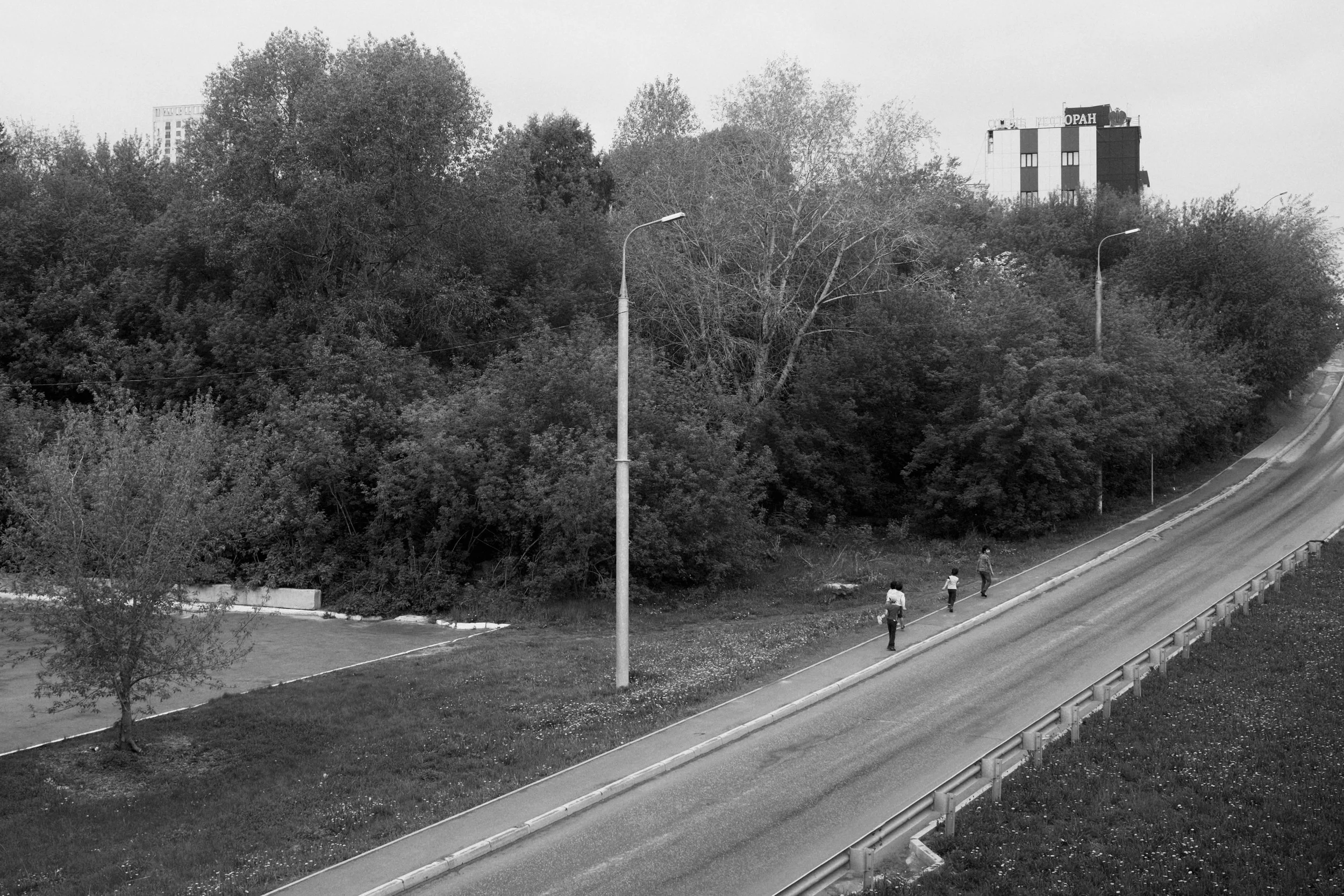 A black and white photo of a sidewalk alongside a road with three people walking, trees behind them, and a tall building in the background.
