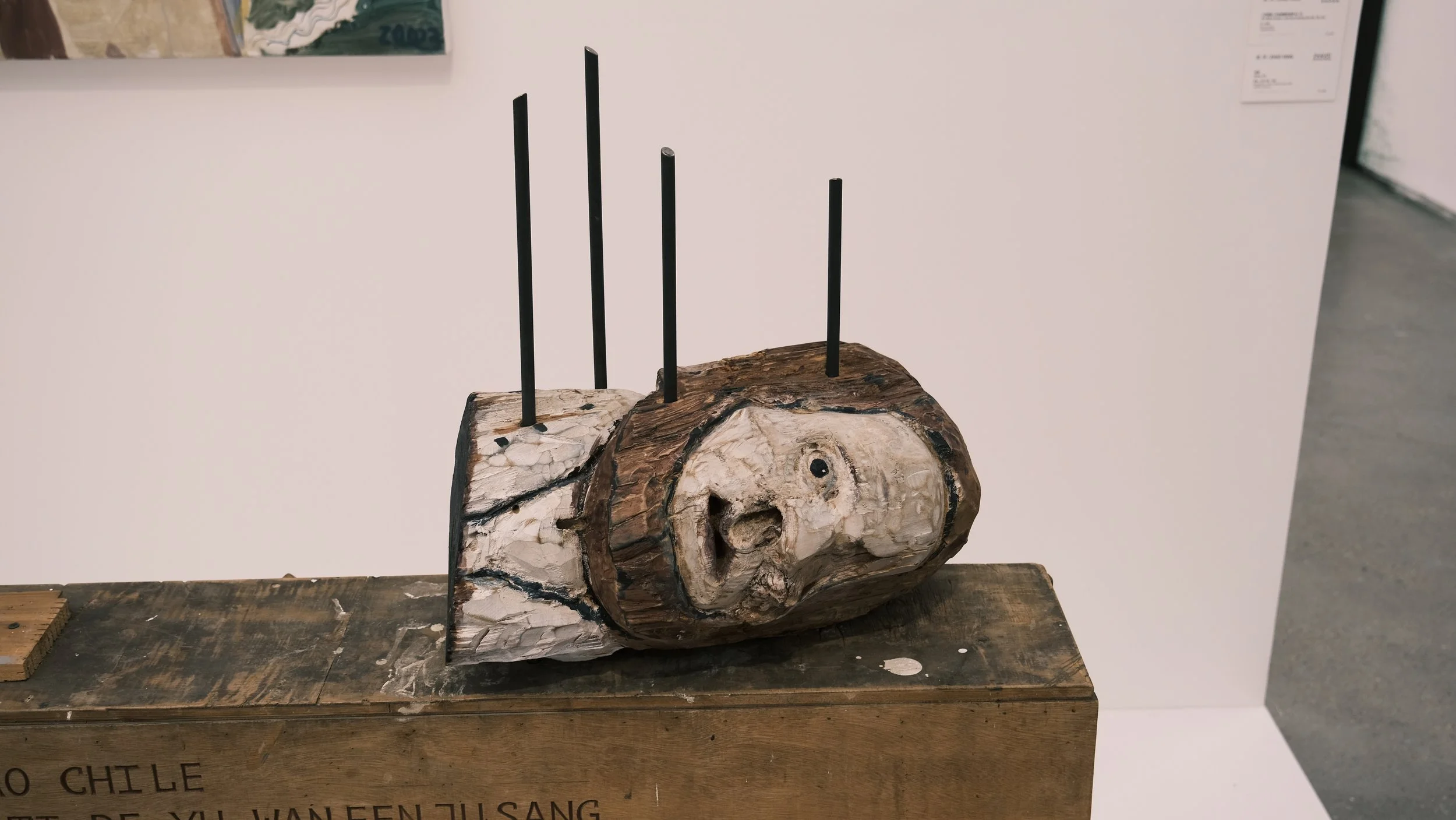 Art sculpture of a head carved into a wood log with black sticks protruding from it, displayed on a wooden surface.