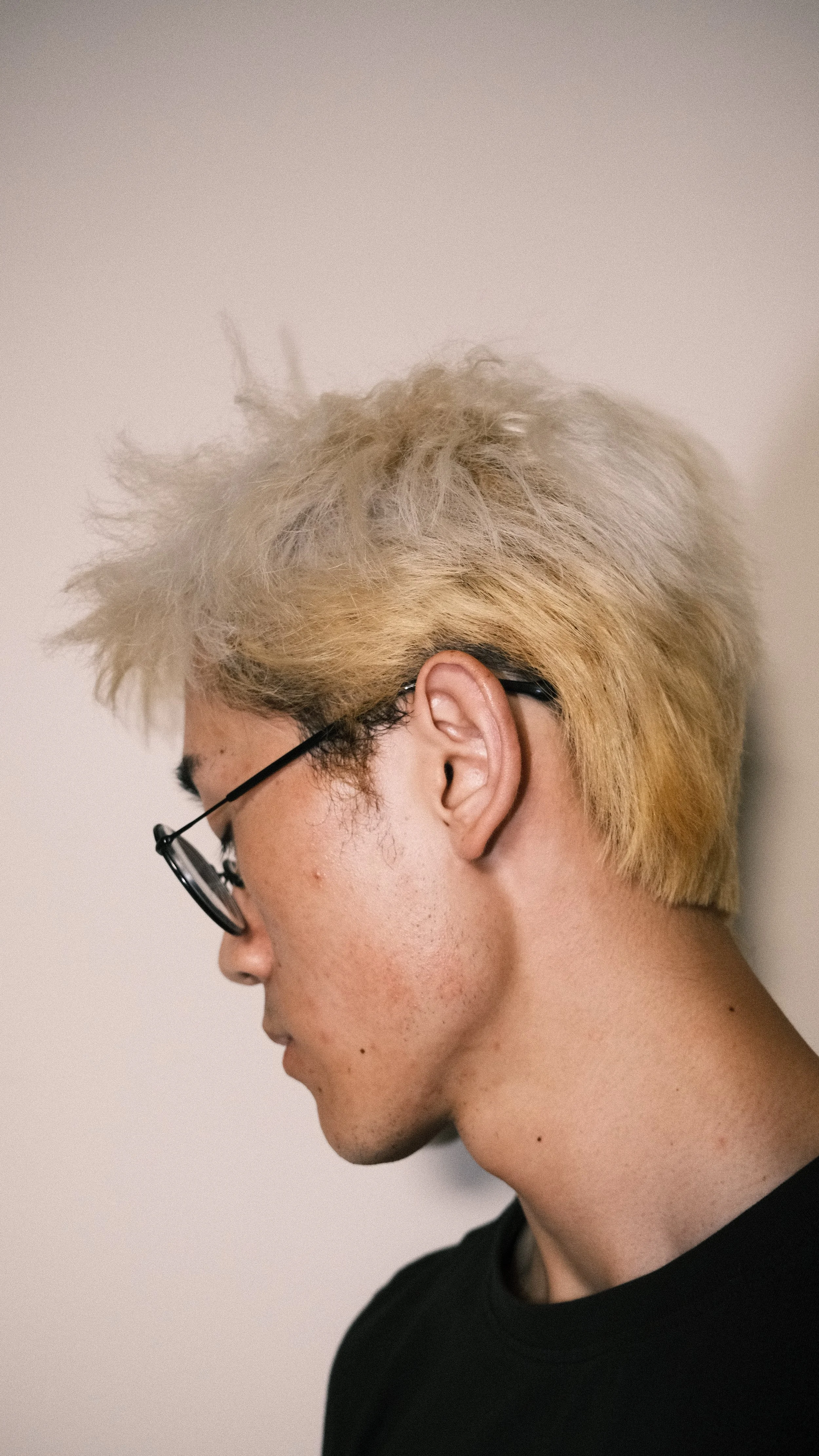 A person with bleached blonde, messy hairstyle wearing glasses and a black shirt, standing against a plain light-colored wall.