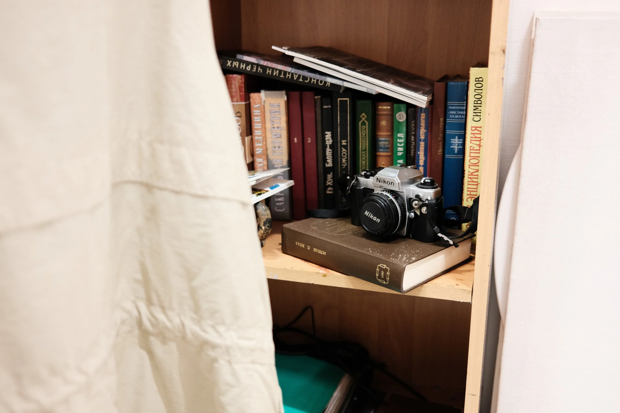 A wooden bookshelf with various books and a black Nikon camera resting on a closed book. Part of a white curtain is visible on the left side of the image.