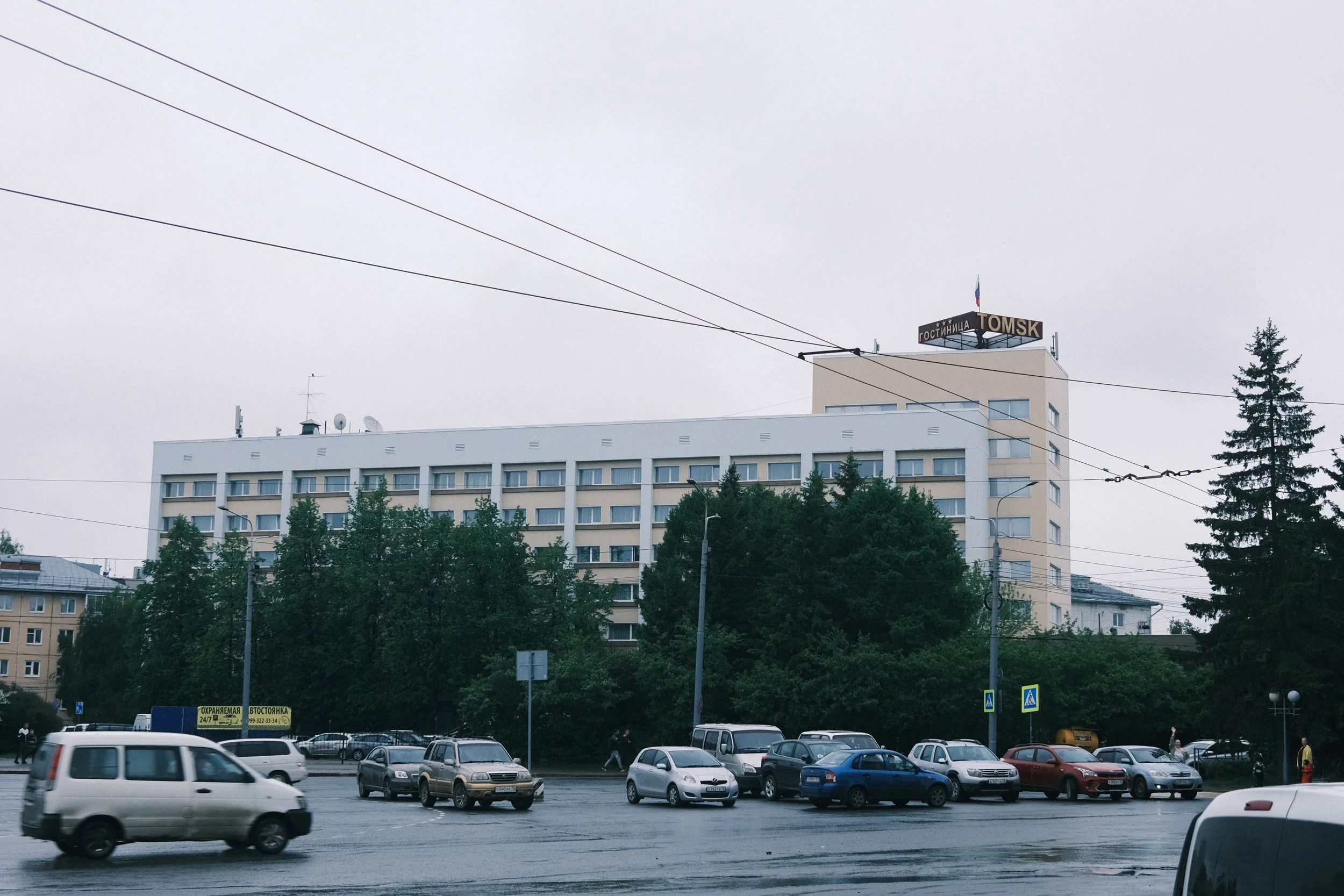 City street scene with cars parked along the curb, a white multi-story building in the background, trees lining the street, overcast sky, city traffic, and a sign on the building reading 'TOMSK' in English and Russian.