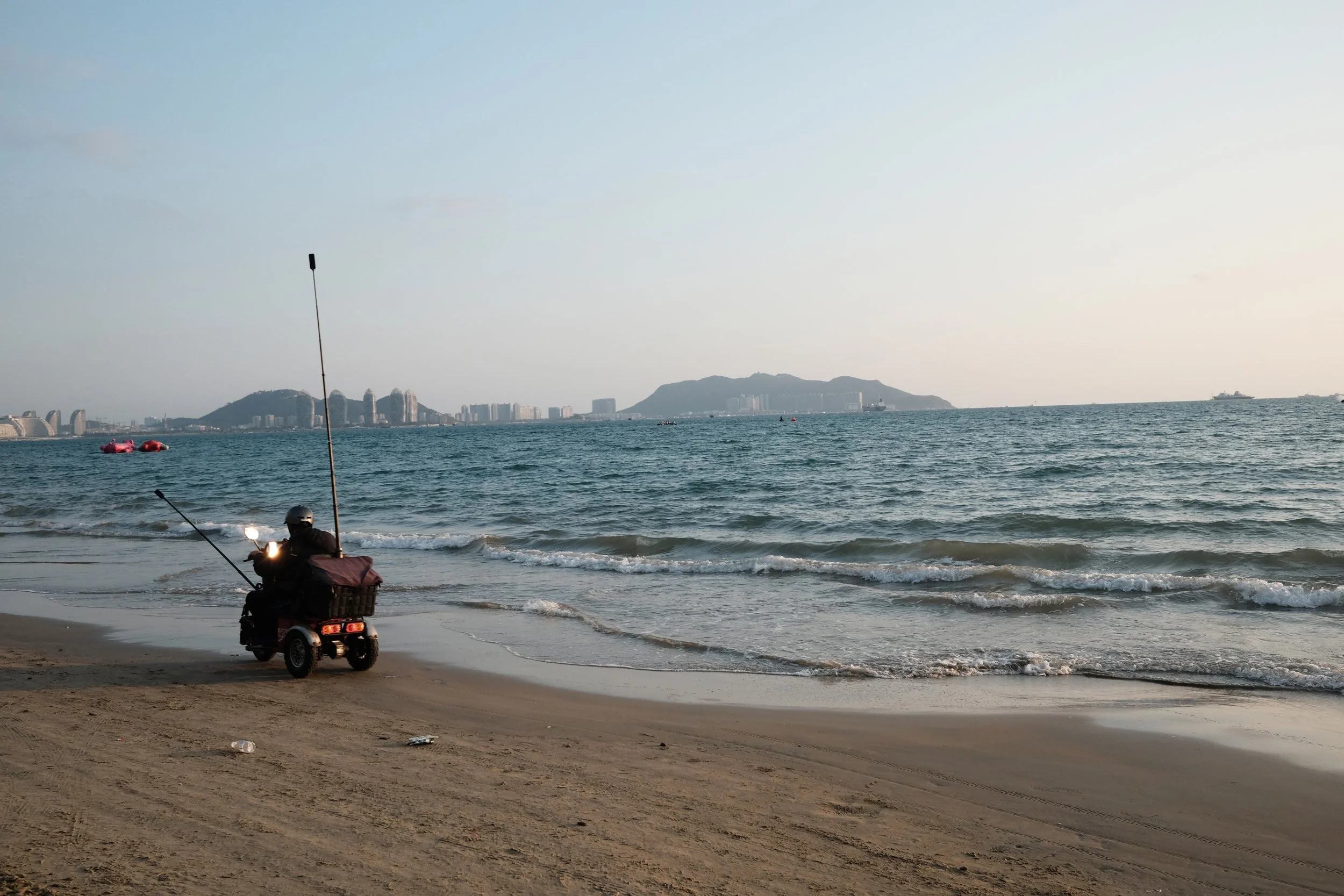 A person riding a motorized scooter with fishing poles on a sandy beach, with a city skyline and mountains in the background under a clear sky.