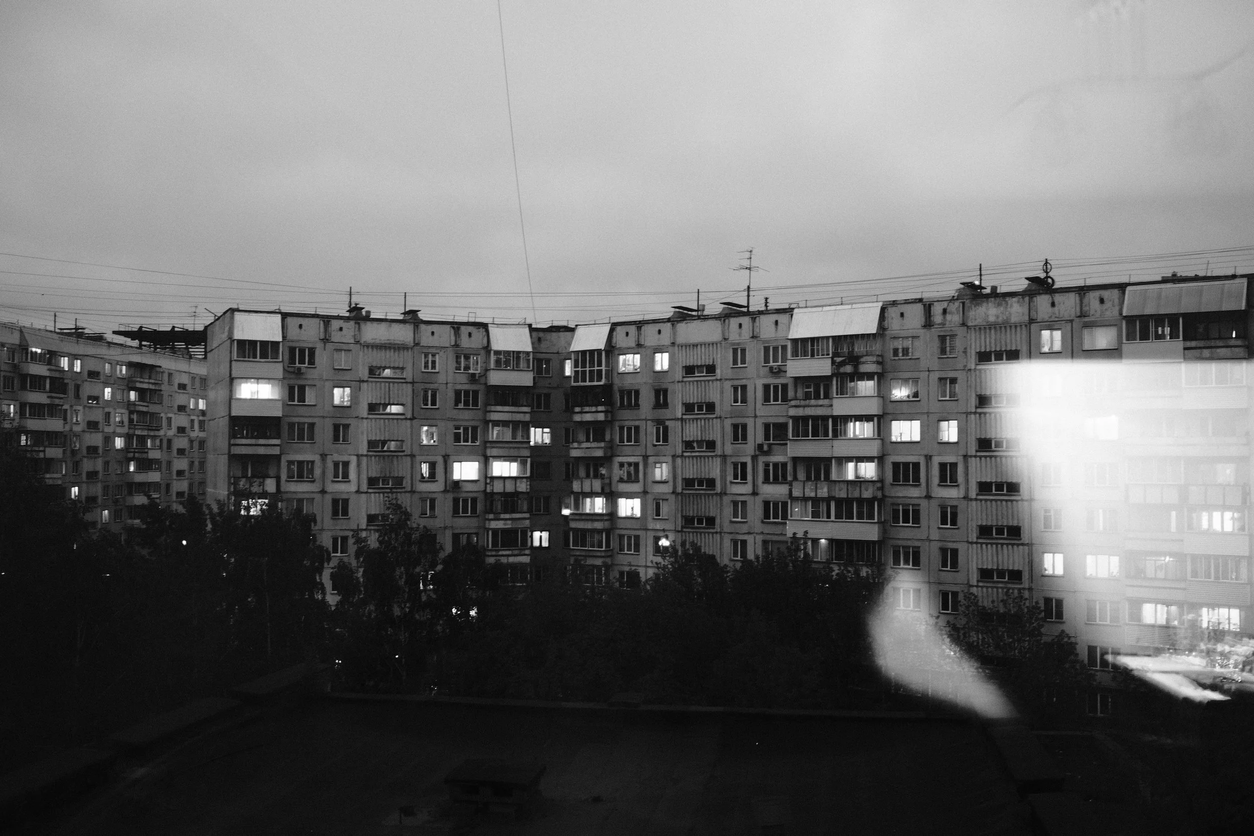 Black and white photo of a large apartment building with multiple windows, some of which have lights on, under an overcast sky at dusk.
