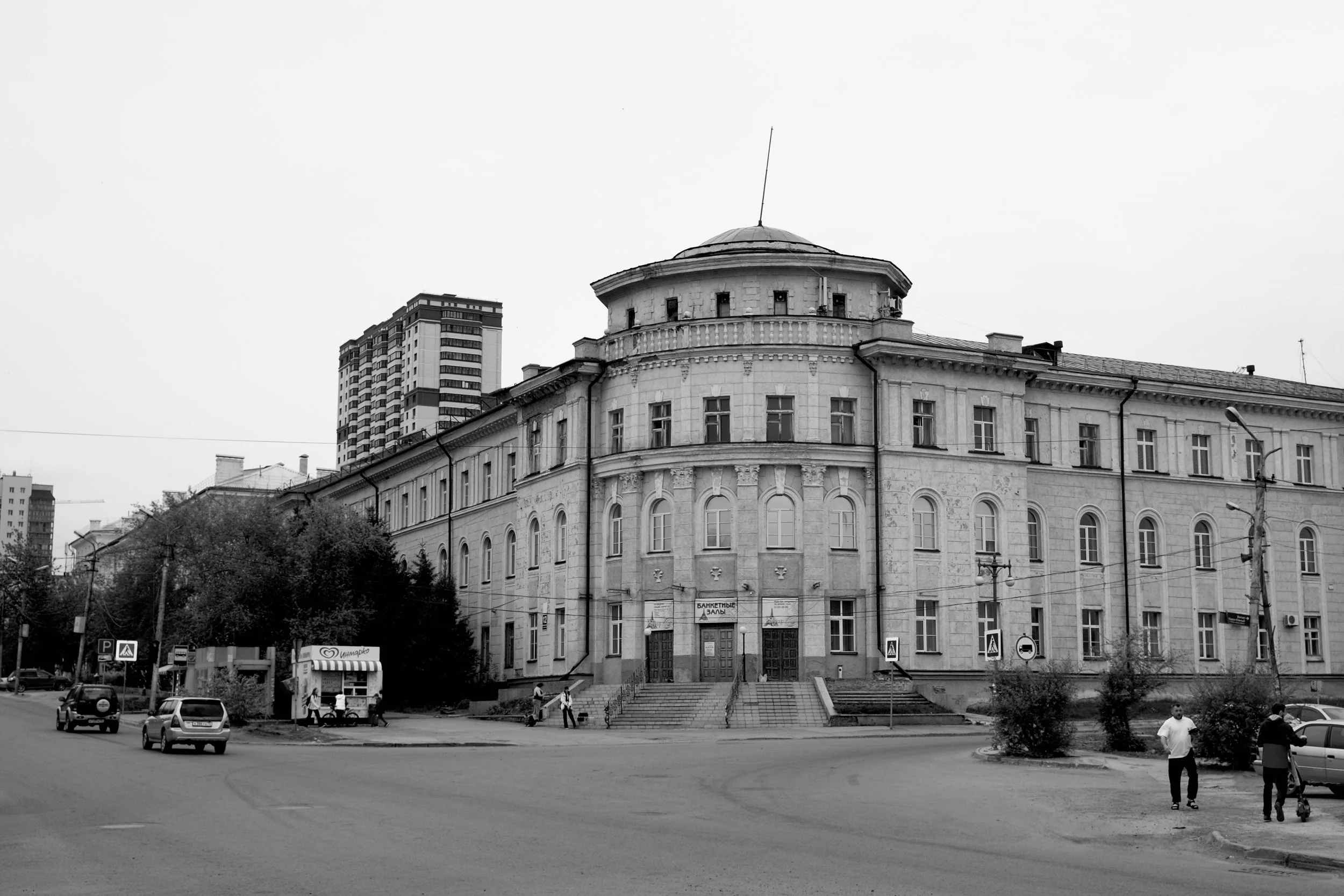 A large historic building with a rounded tower, stairs leading to the entrance, and several windows, situated on a street with a few cars and pedestrians, black and white photograph.